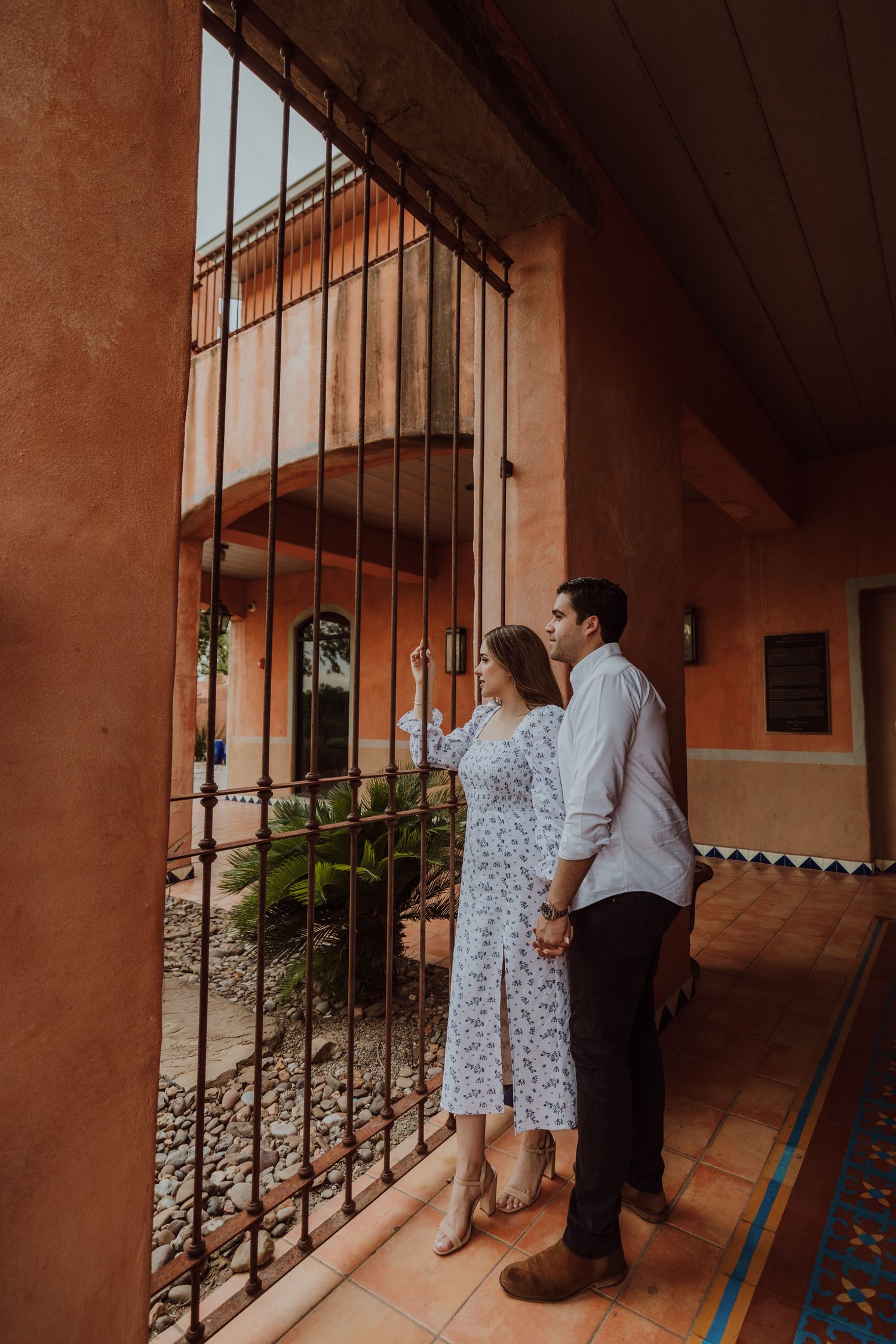 Couple holding hands exploring a Spanish colonial building with ornate iron gate and terracotta architecture.