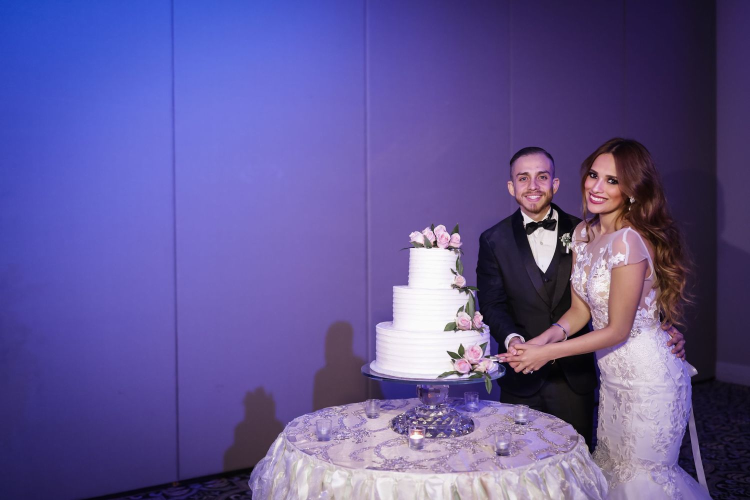 Couple in formal wear smiling while cutting tiered white wedding cake at reception.