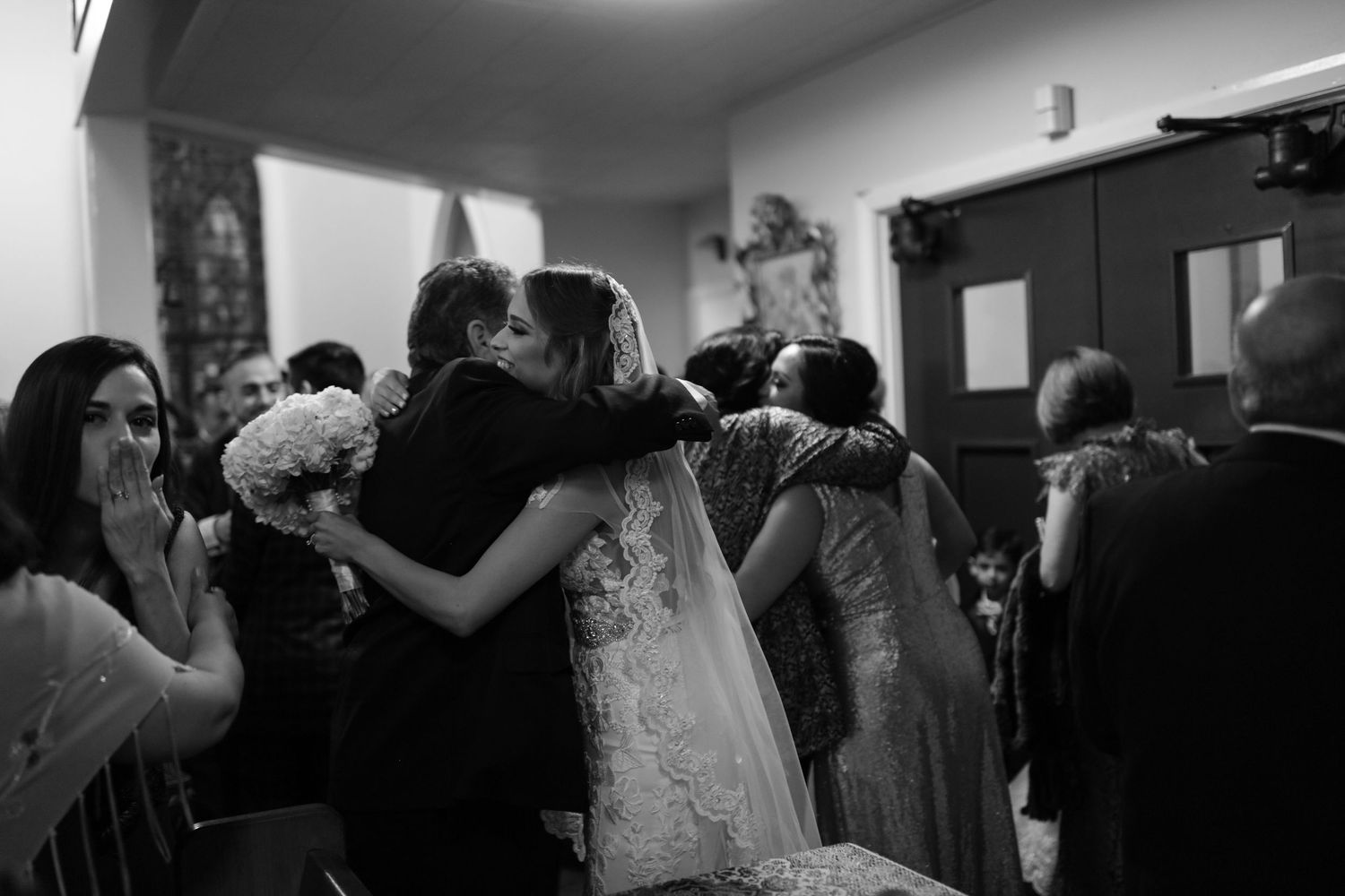 Bride in lace wedding gown embraces guests in emotional black and white photo inside church after ceremony.