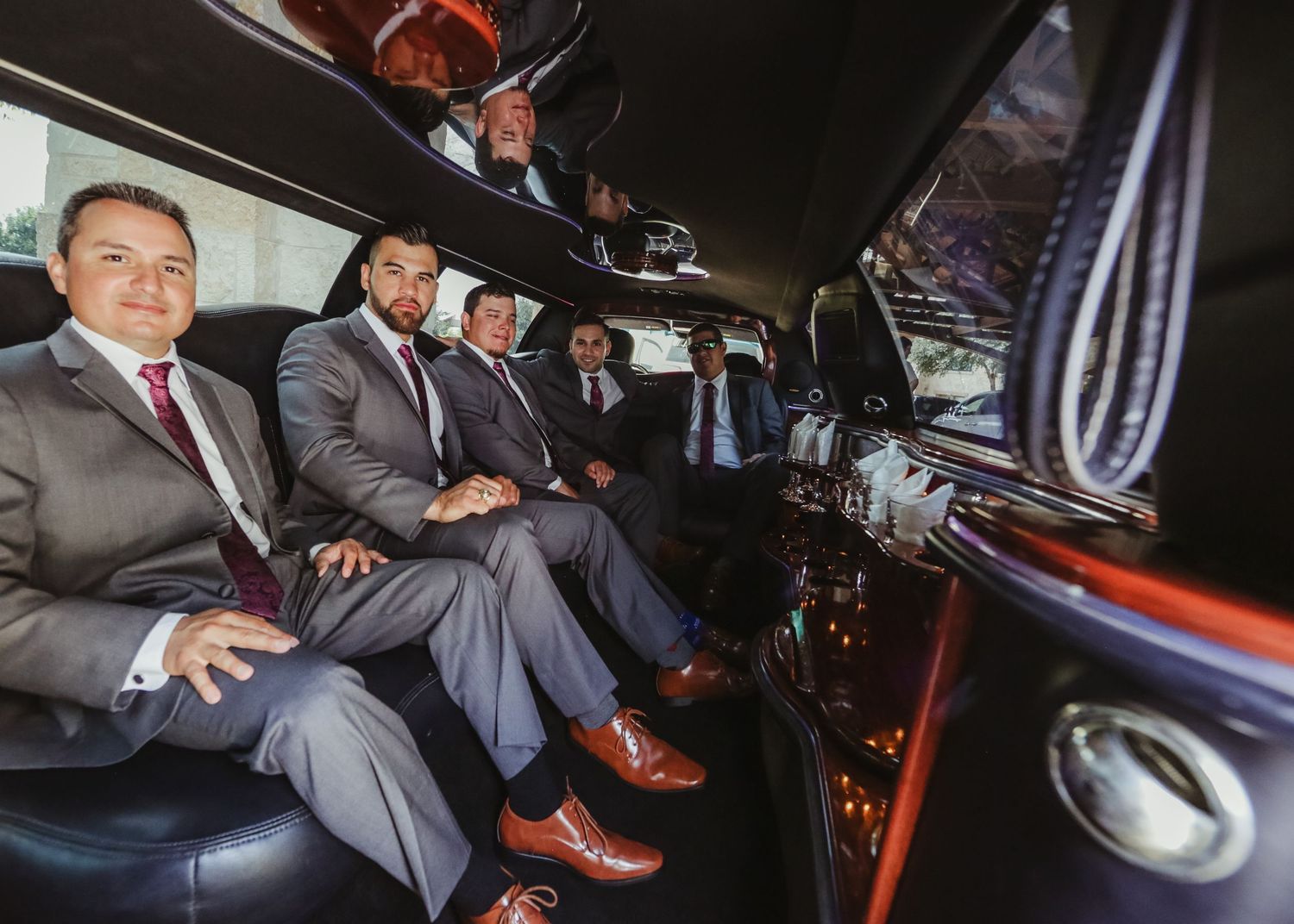 Groomsmen in gray and navy suits laugh together inside a black stretch limousine before the wedding.