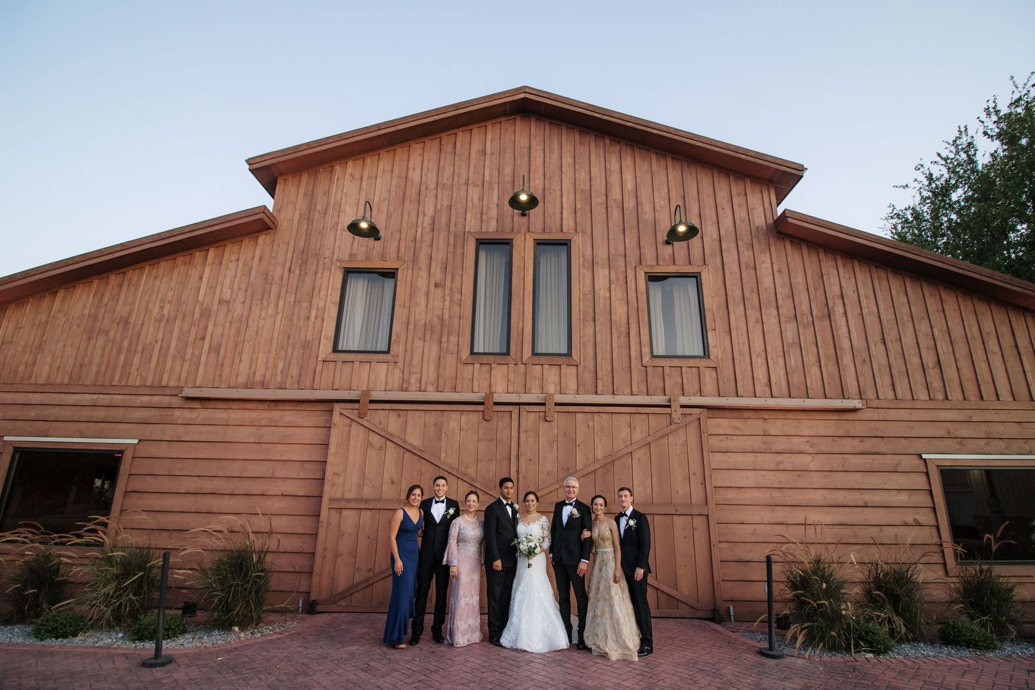 Wedding party poses in front of rustic red barn venue at dusk.