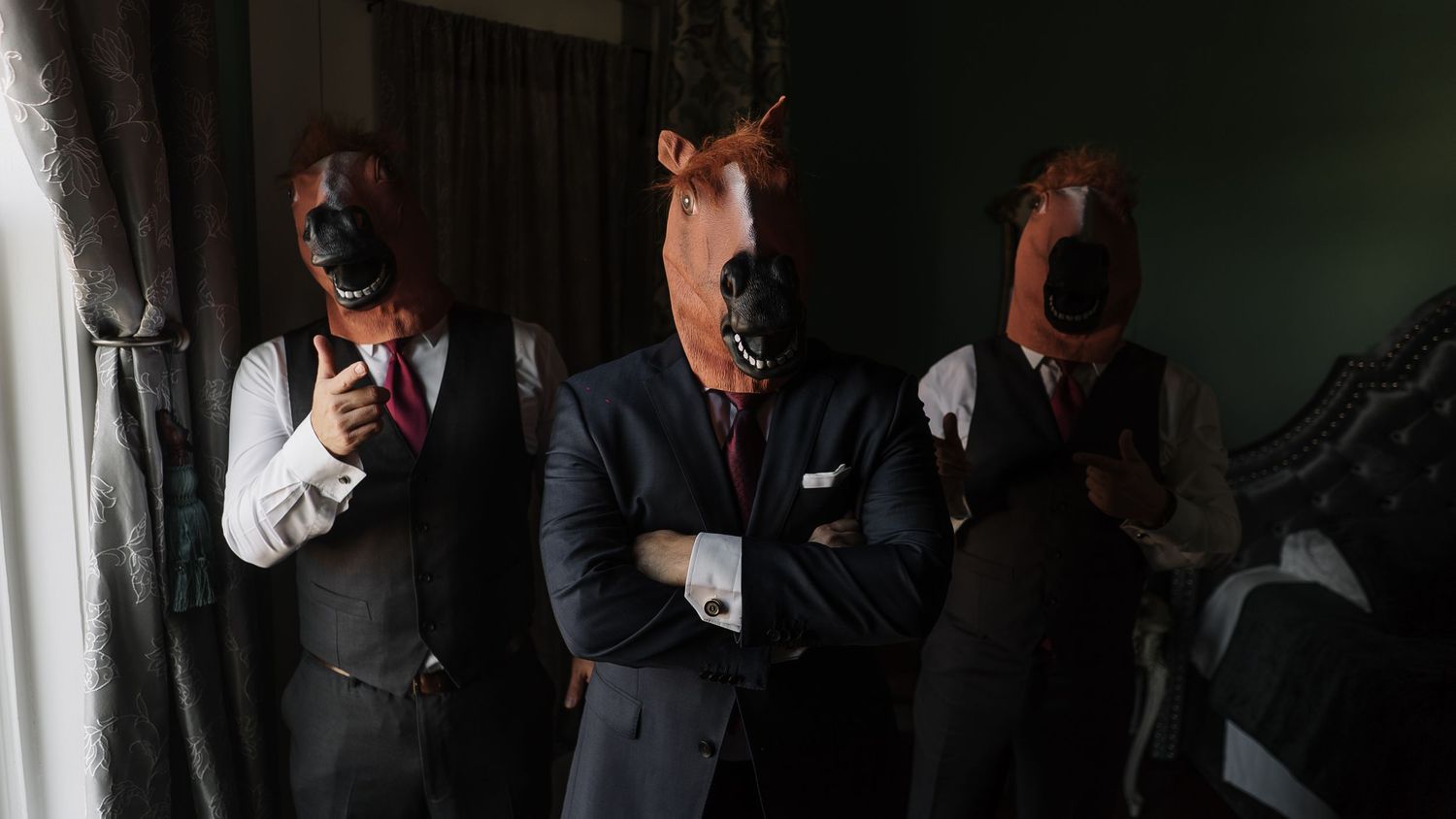 Three men in suits wearing horse and black masks pose formally indoors in a dramatic, mysterious setting.