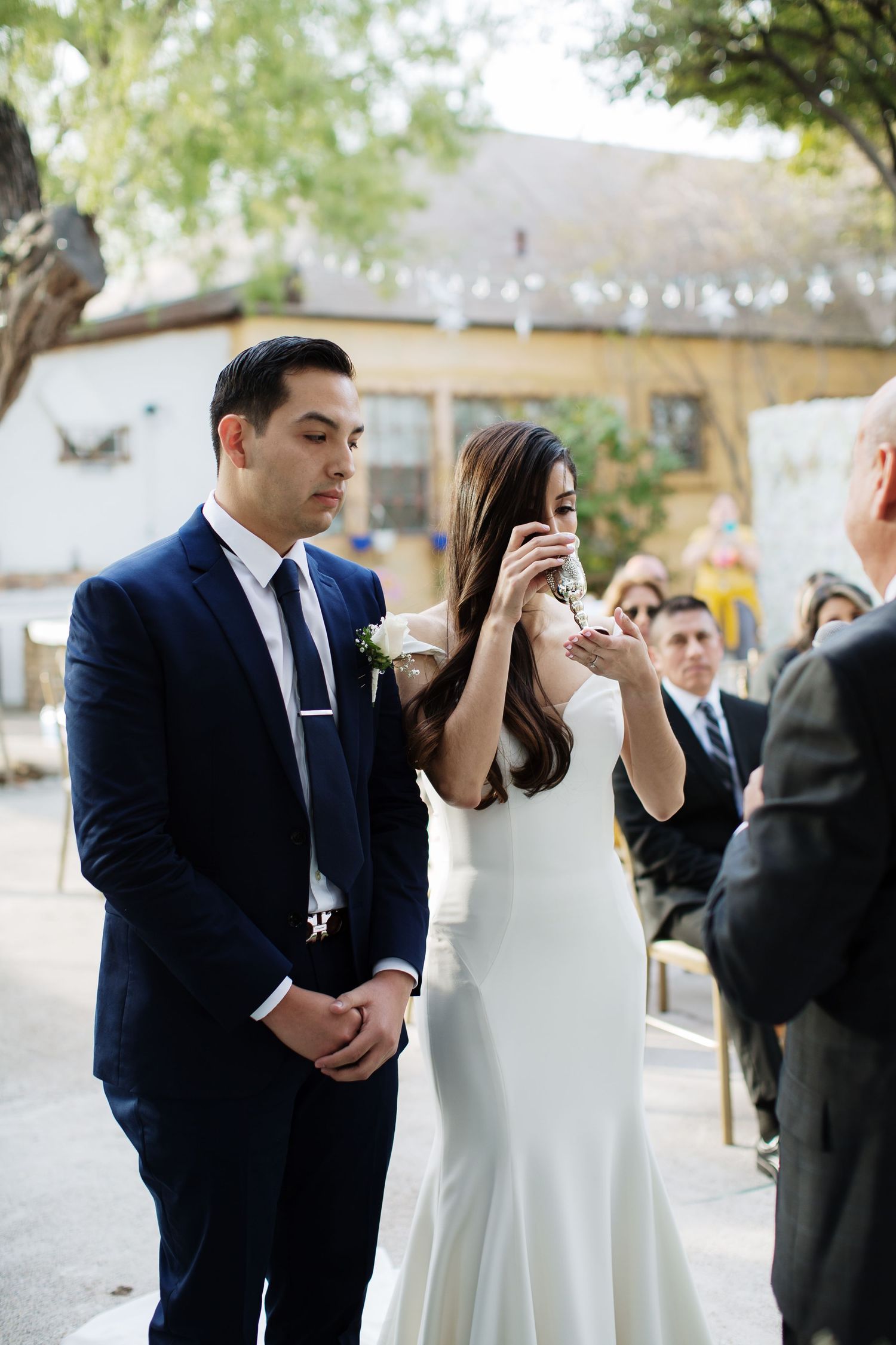 A bride wipes tears during an outdoor wedding ceremony beside her groom in a navy suit.