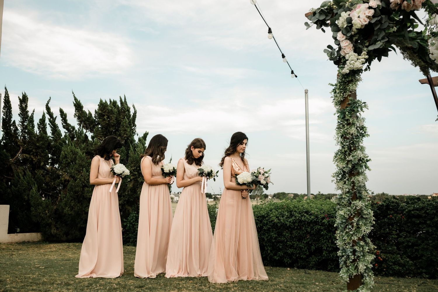 Four bridesmaids in blush pink gowns stand near floral wedding arch outdoors.