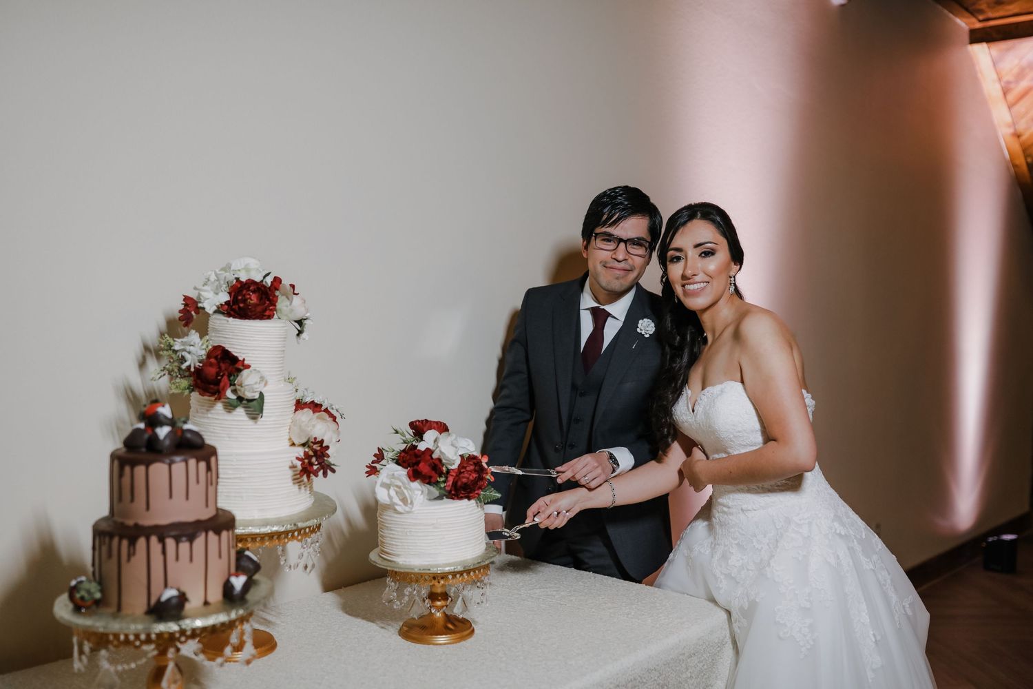 Bride and groom smile while cutting tiered white wedding cake with red floral decorations.