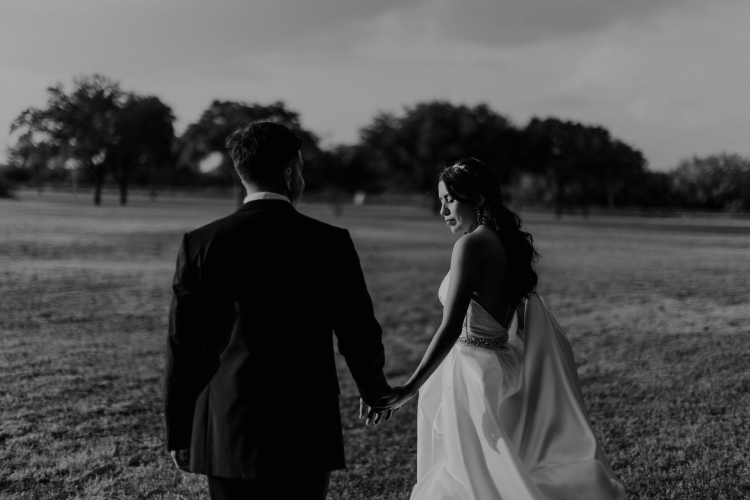 Black and white photo of a bride and groom holding hands walking through a field on their wedding day.