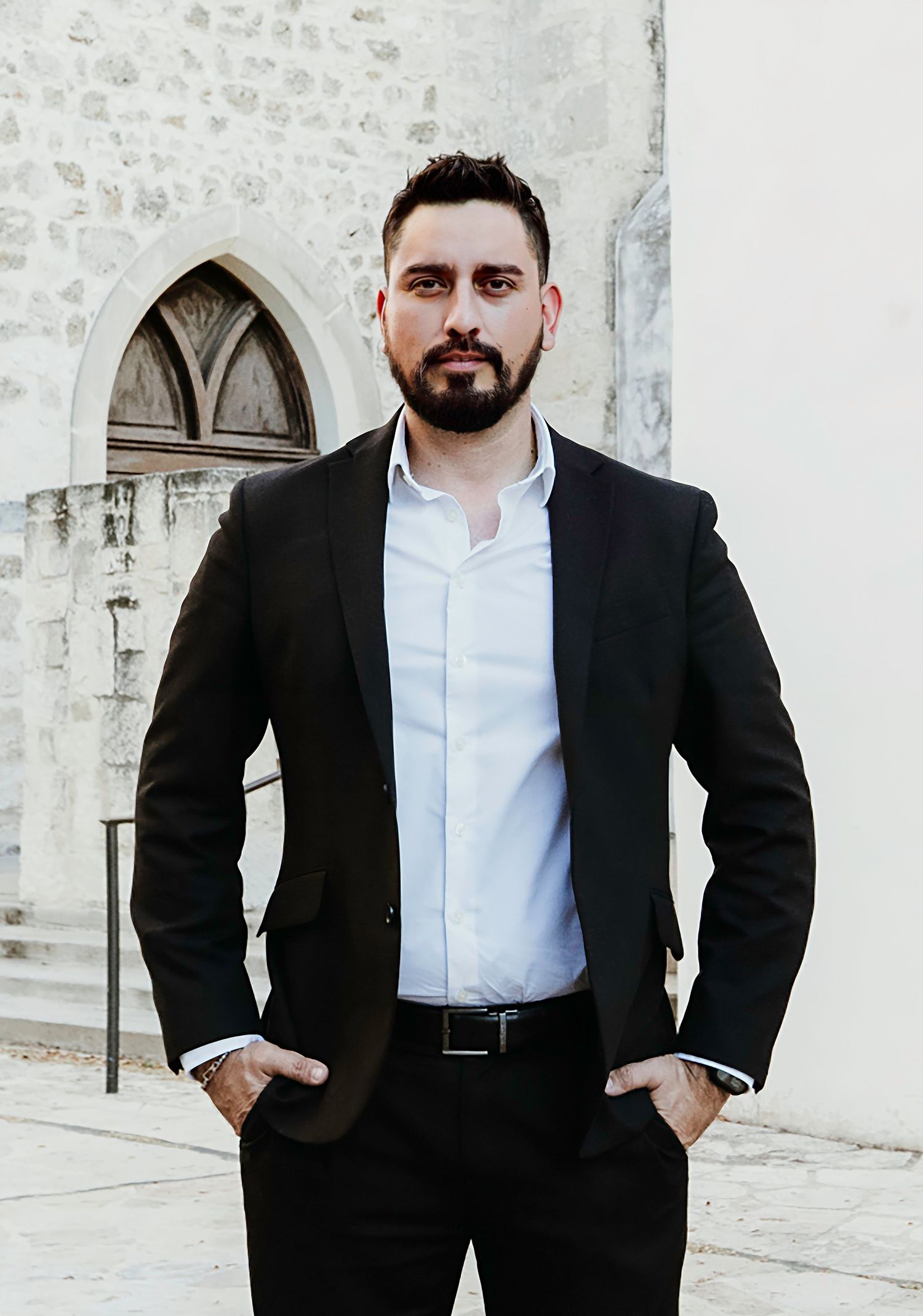 Professional standing against white stone wall with arched gothic window wearing black suit and white shirt.