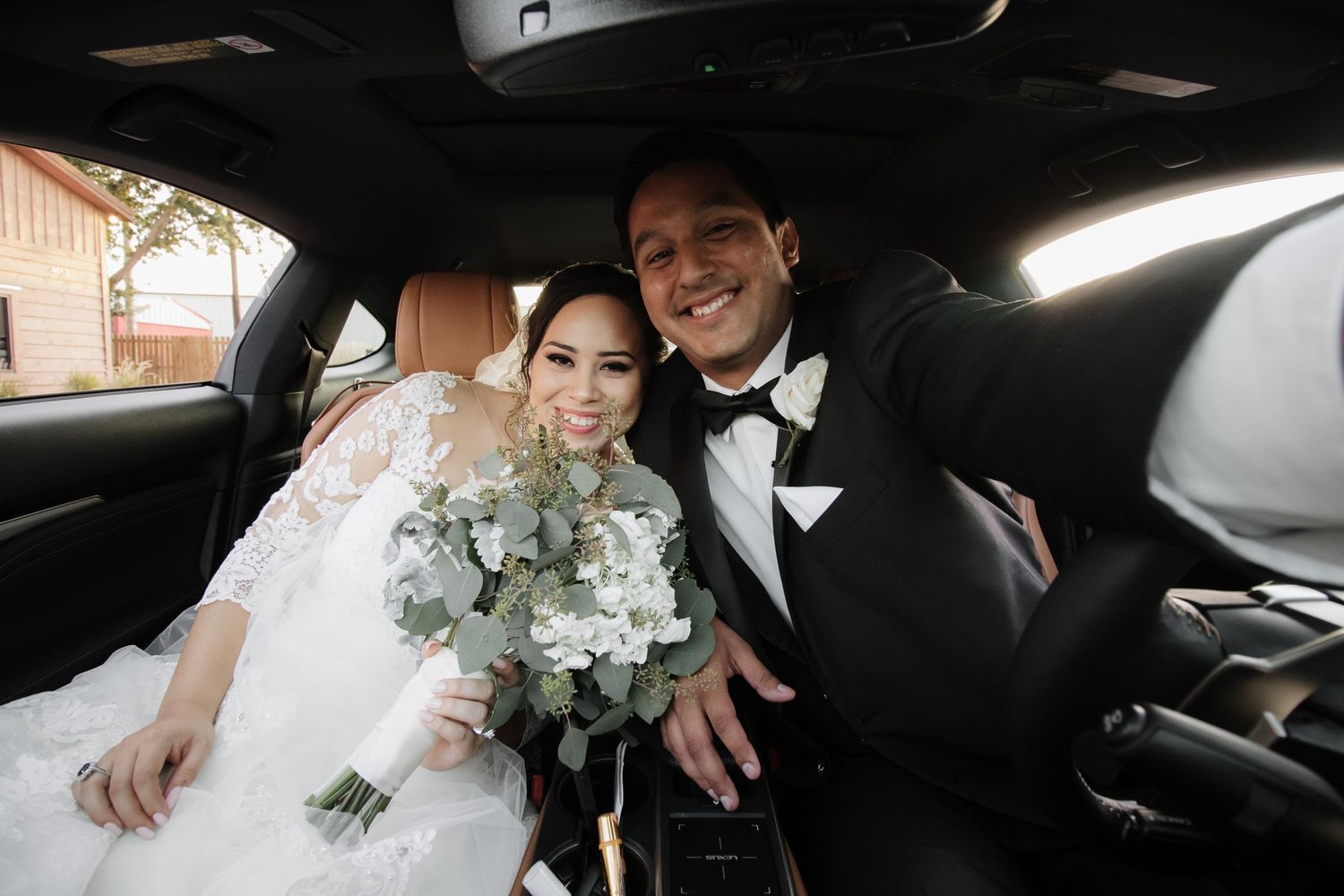 Happy newlyweds smiling together in back seat of car with bridal bouquet.