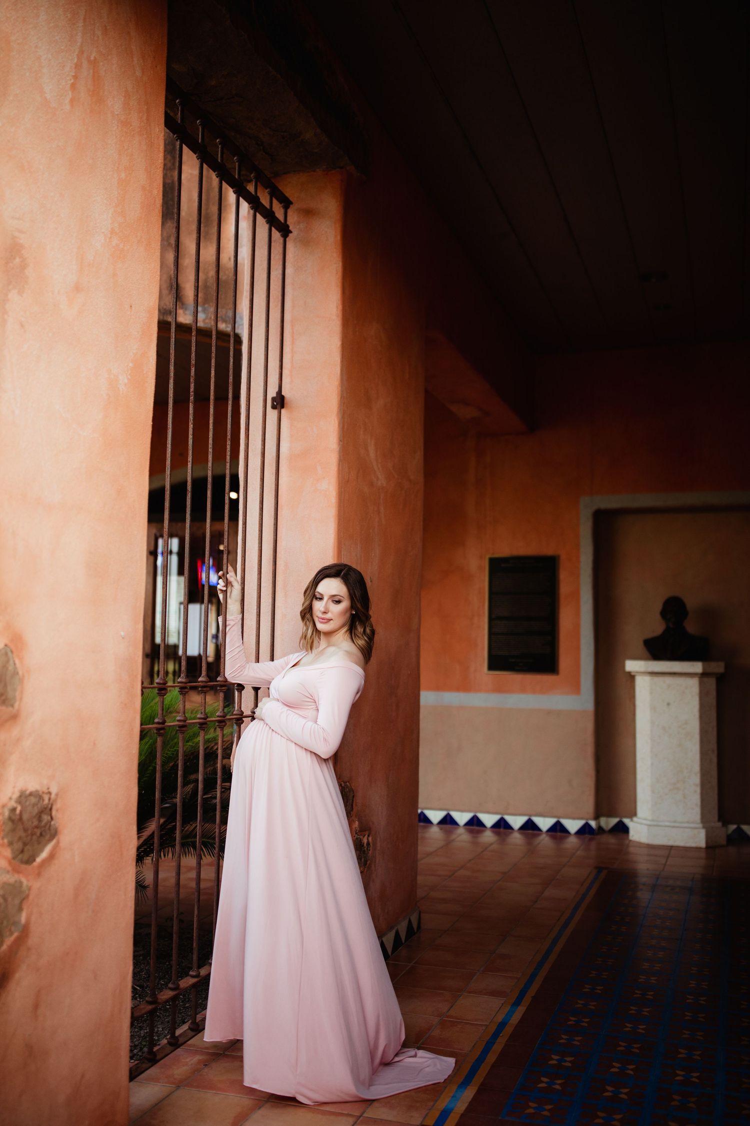 Elegant maternity portrait in a pink dress beside ornate iron gates and peach-colored Spanish colonial walls.
