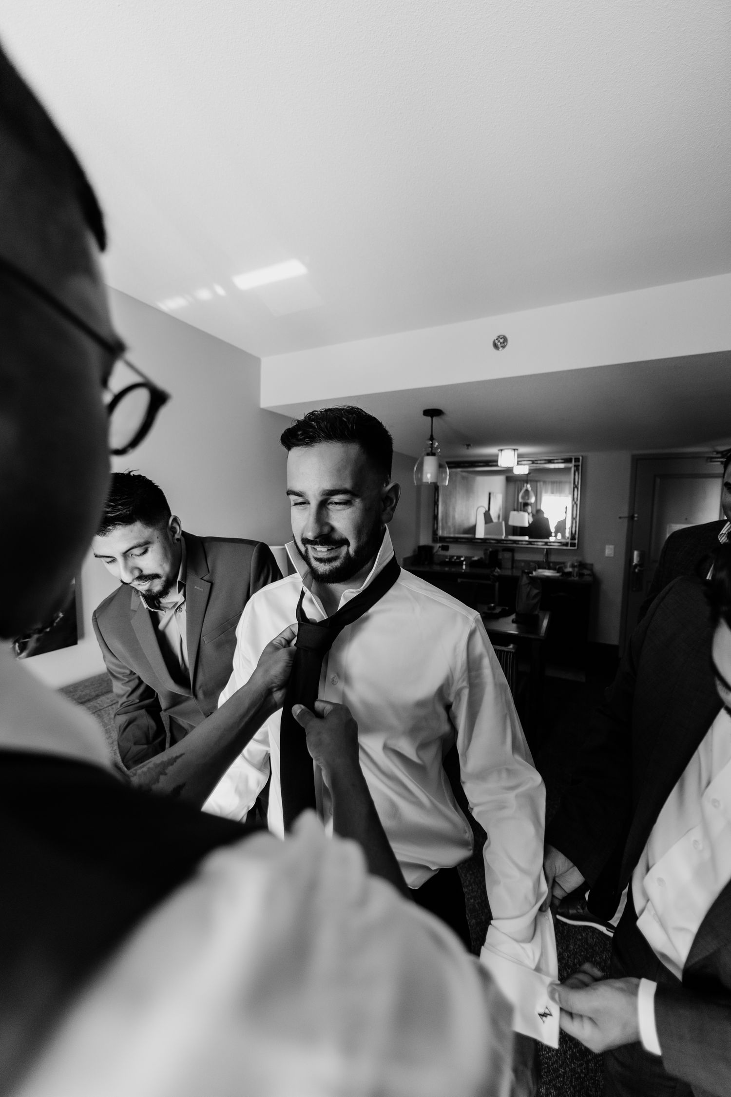 Black and white photo of groomsmen helping groom adjust his tie while getting ready for wedding day.