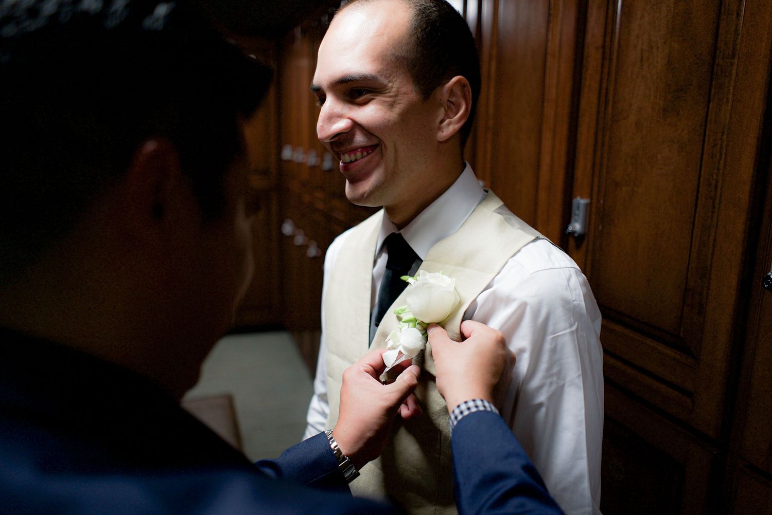 Smiling groom having a white boutonniere pinned to his vest by a groomsman before the wedding.