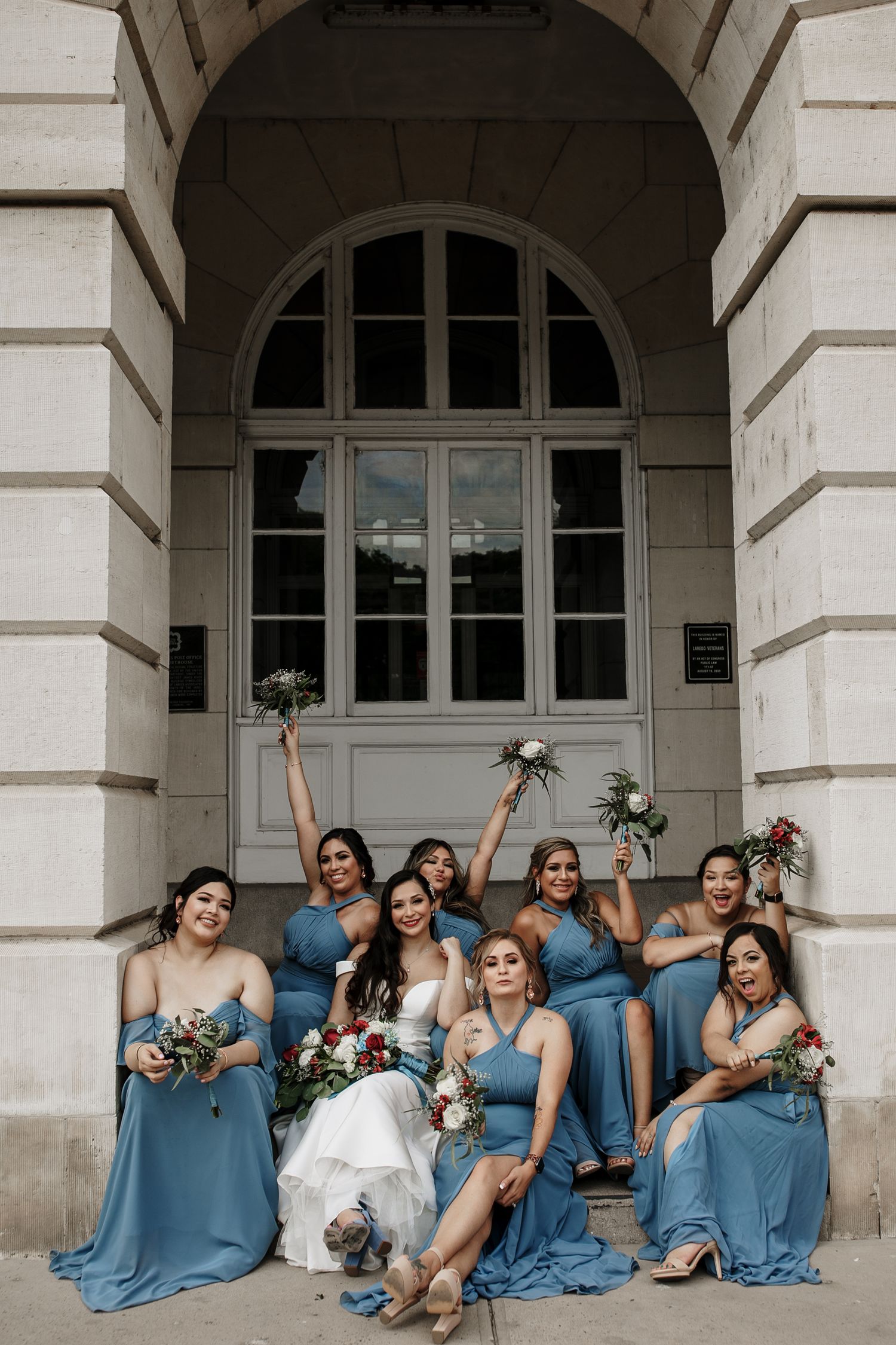 Bride in white gown celebrates with bridesmaids in blue dresses raising bouquets on steps of grand building.