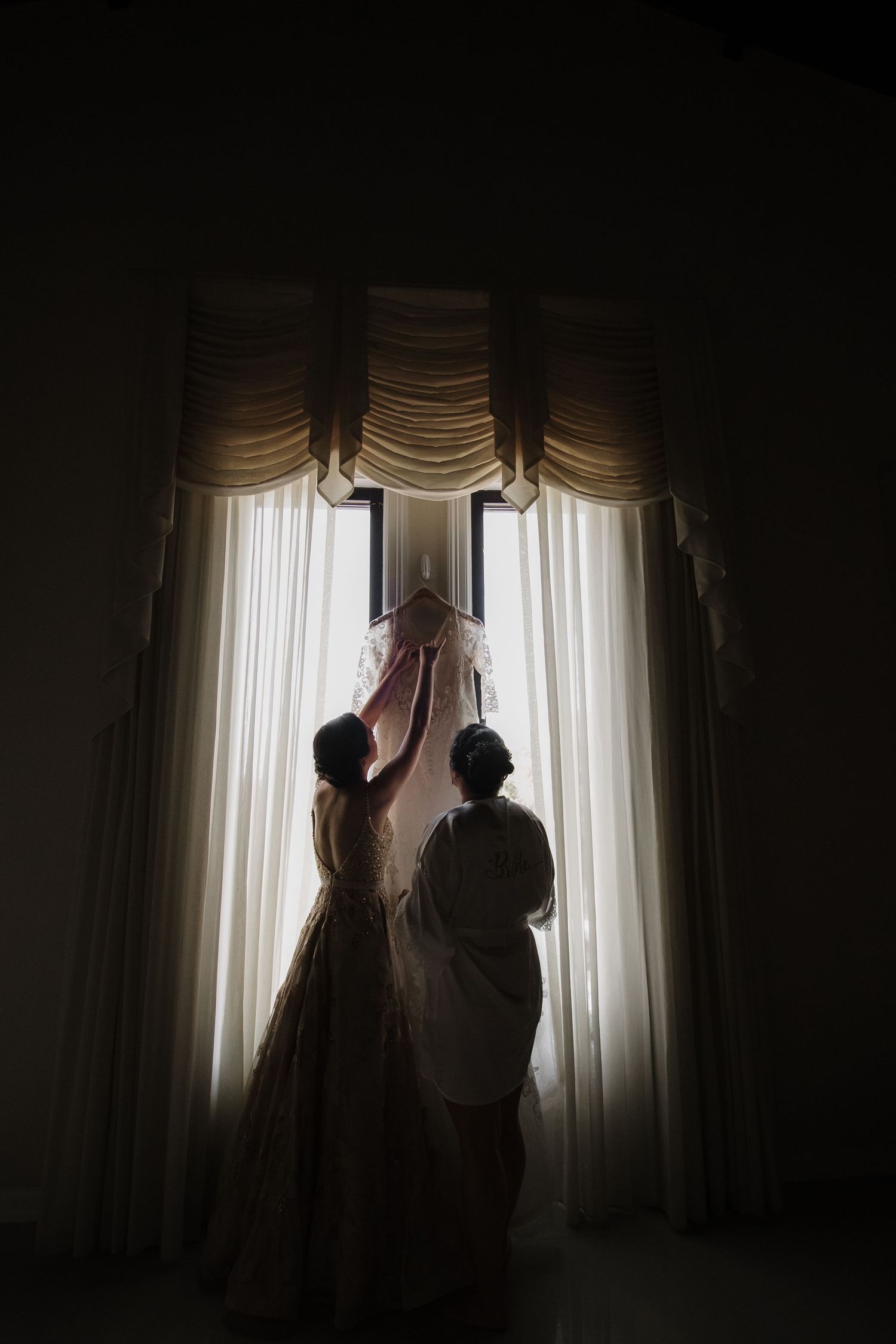 Silhouette of two women holding up a wedding veil in front of a draped window.