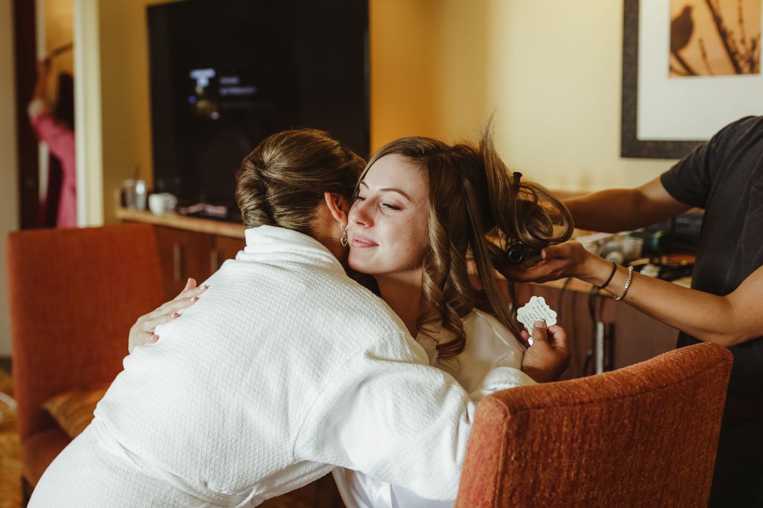 Emotional bride receiving a hug while getting hair styled in a white robe before her wedding.