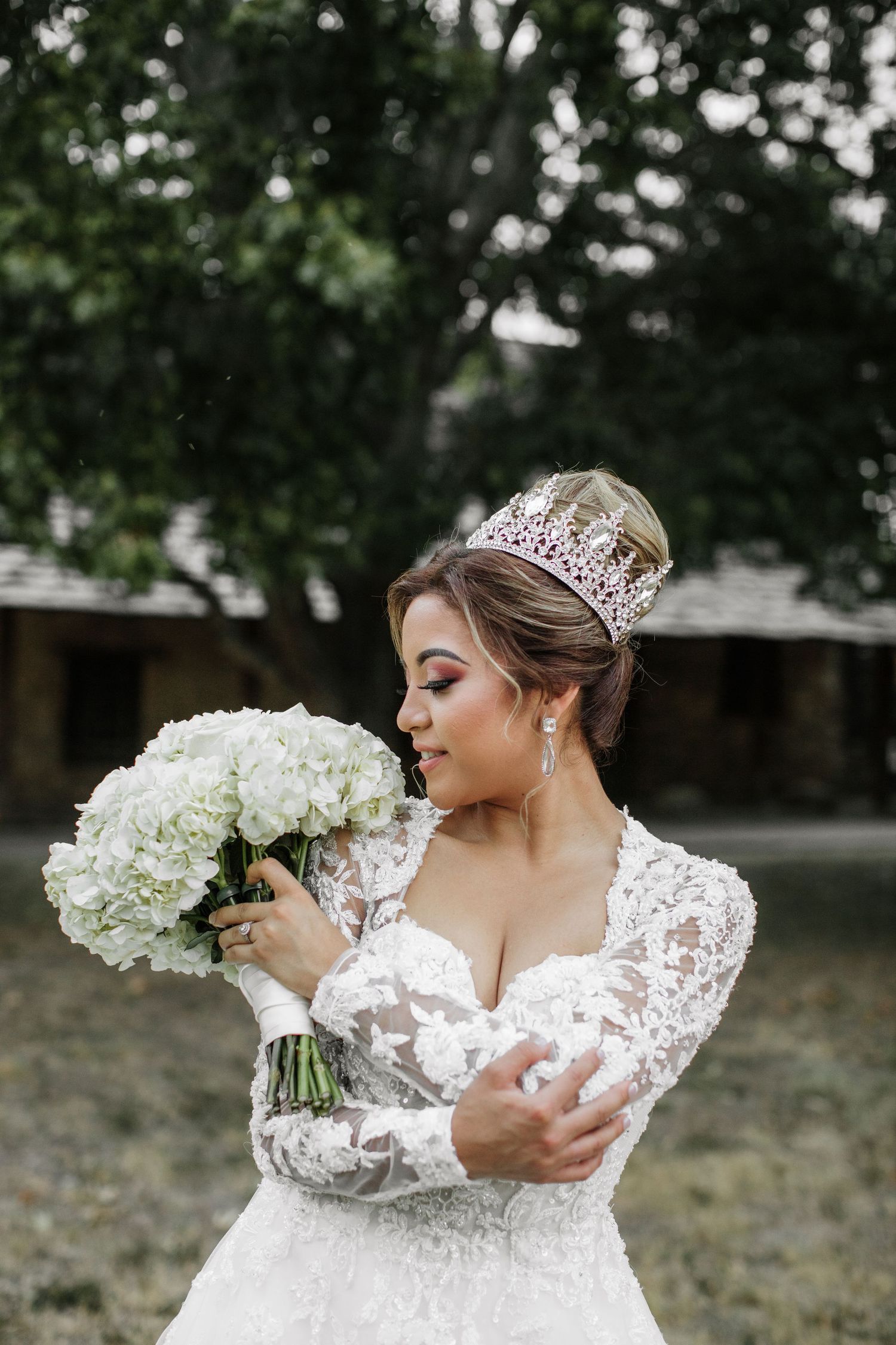 Bride wearing lace gown and crystal crown holds white hydrangea bouquet outdoors.