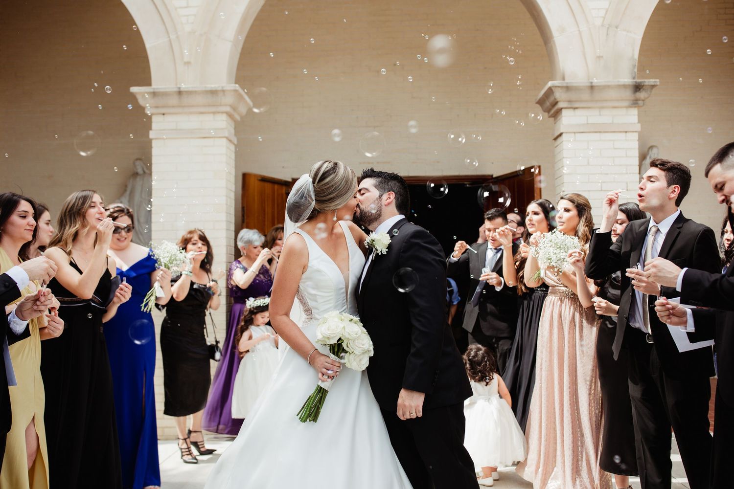Newlyweds share a kiss as guests toss confetti outside elegant arched wedding venue.