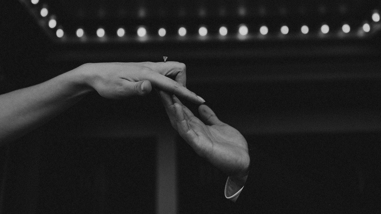 Black and white image of hands touching under string lights at night.