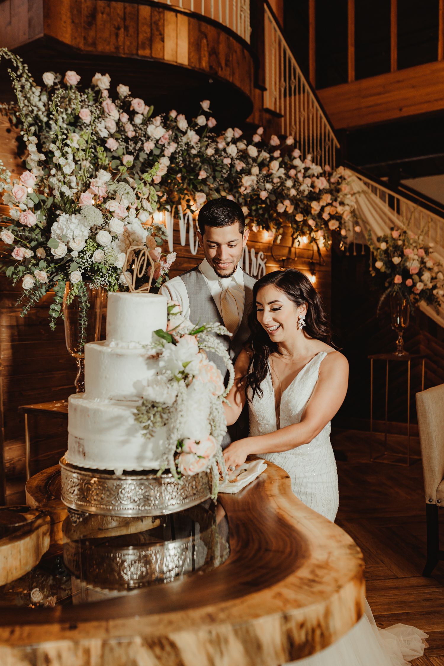 Bride and groom smiling while cutting a white floral wedding cake on a wooden slab table in a rustic venue.
