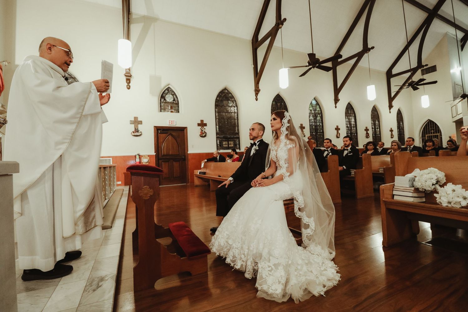 Bride and groom kneel at the altar during a traditional Catholic wedding ceremony in a white chapel.
