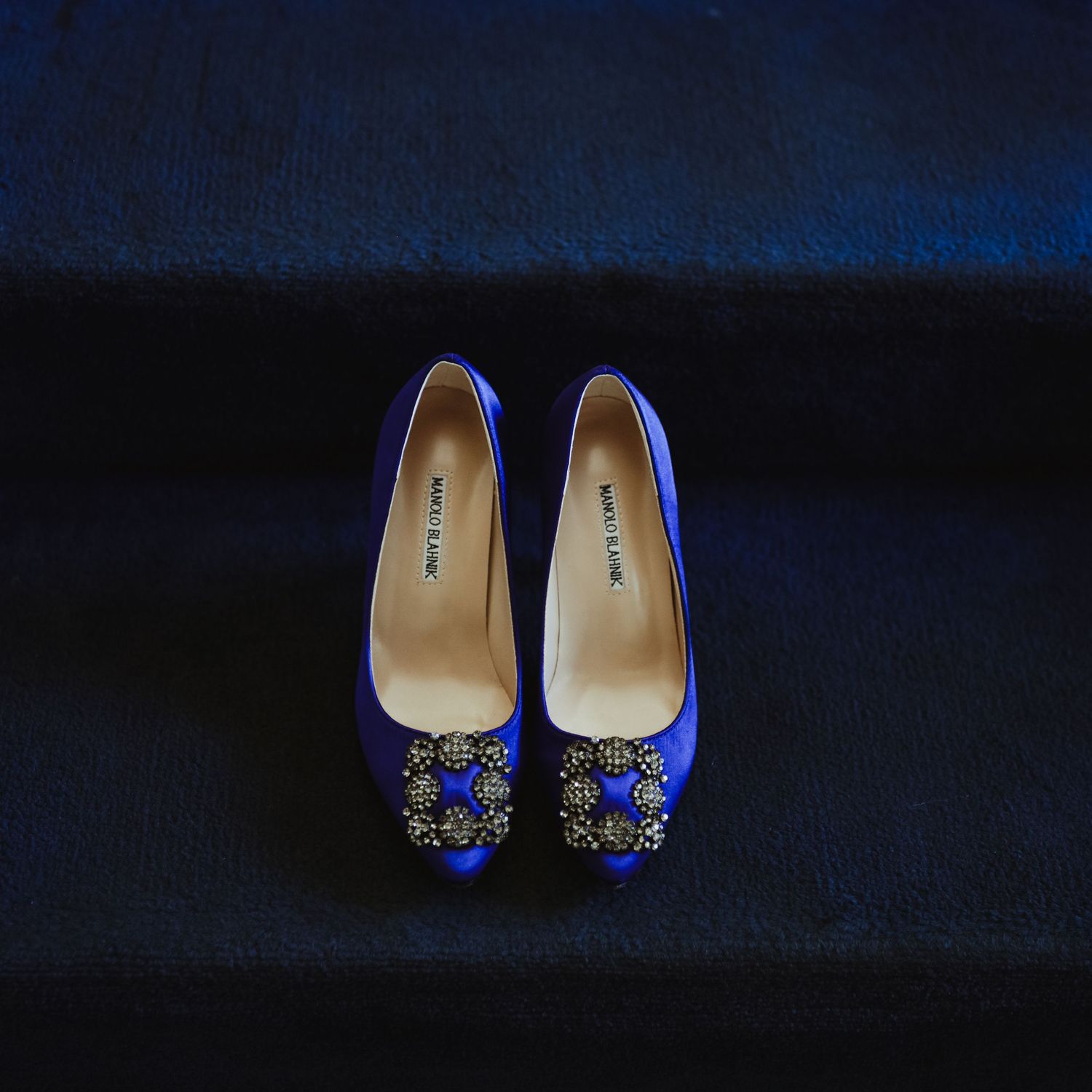 Elegant blue satin Manolo Blahnik heels with jeweled buckle detail on dark velvet surface.