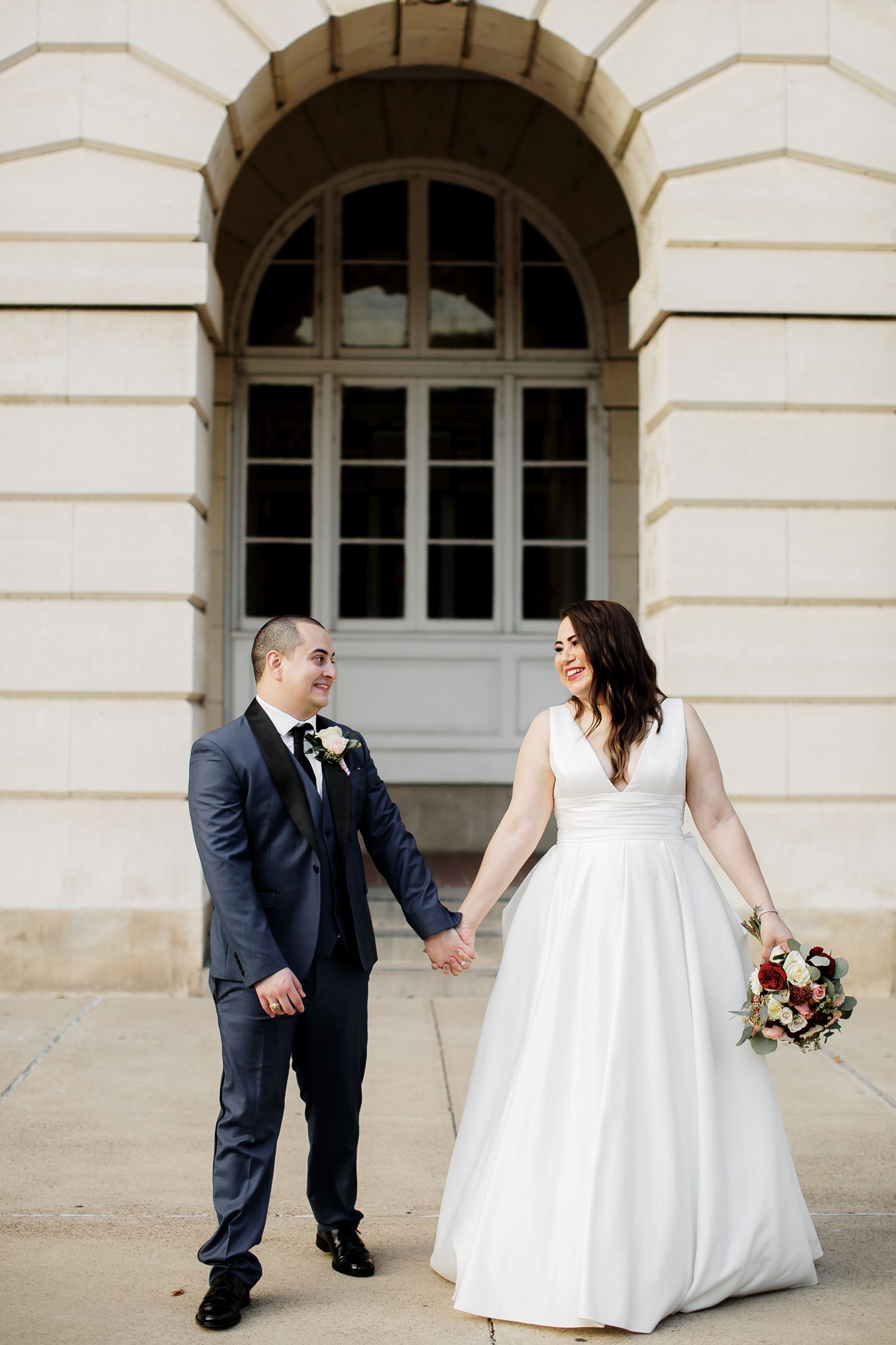 Newlyweds holding hands and smiling while walking outside a grand stone building.