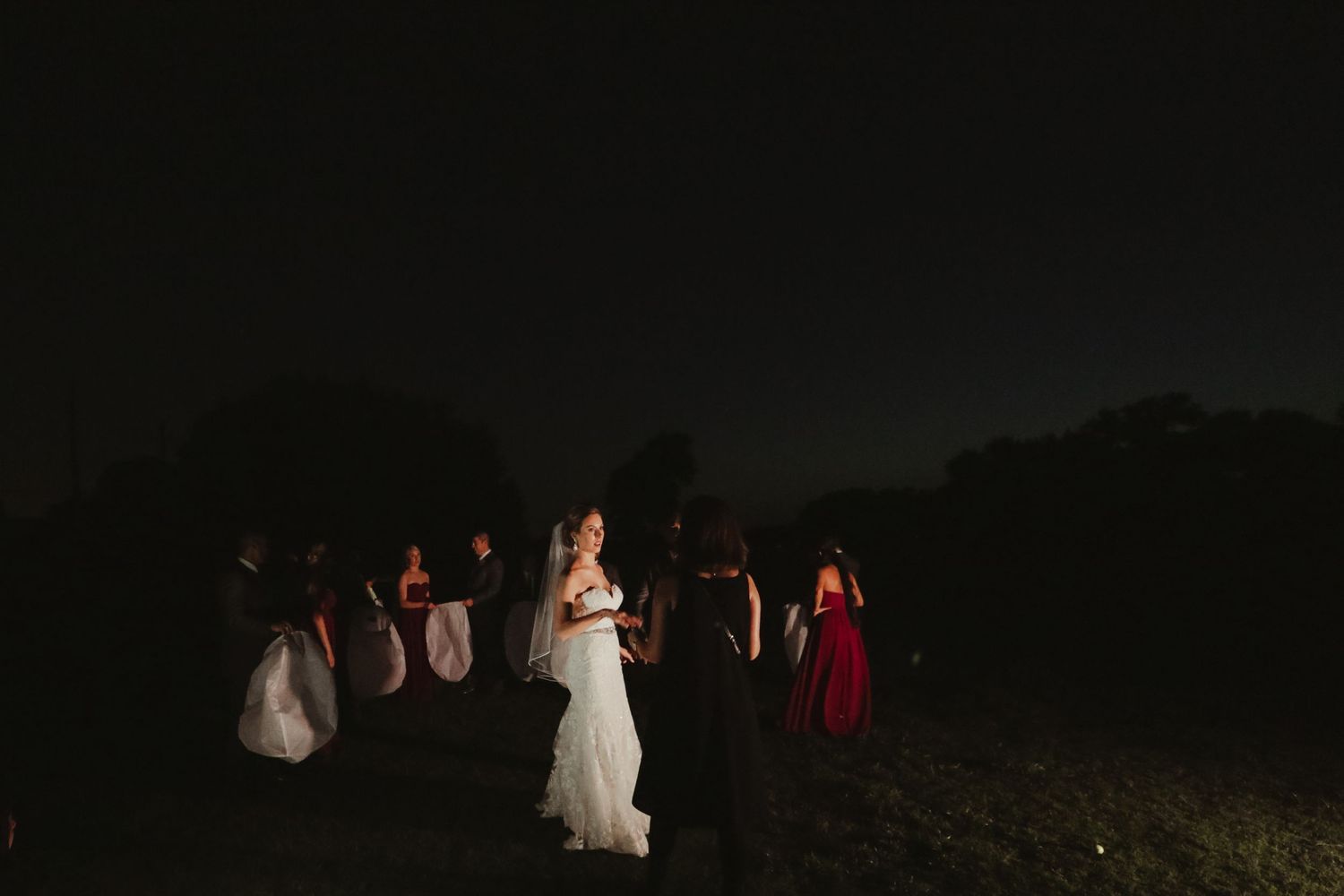 Bride and groom share a quiet moment outdoors at night surrounded by wedding party under dark sky.