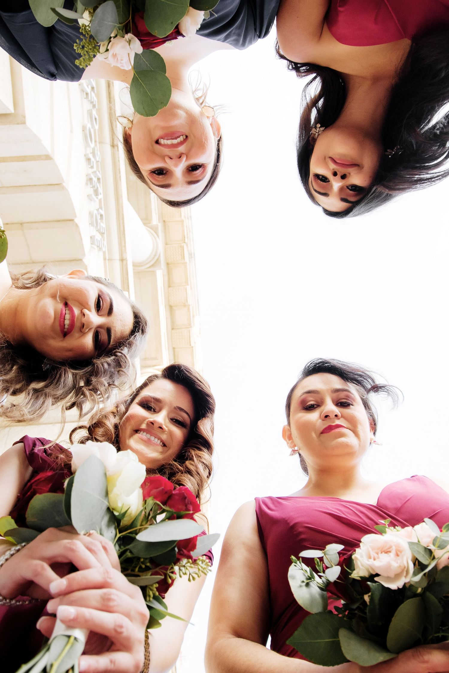 Low angle view of smiling bridesmaids in burgundy dresses holding bouquets of roses and eucalyptus.