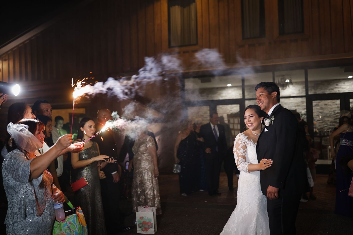Newlyweds walk through sparkler send-off tunnel outside rustic barn wedding venue at night.