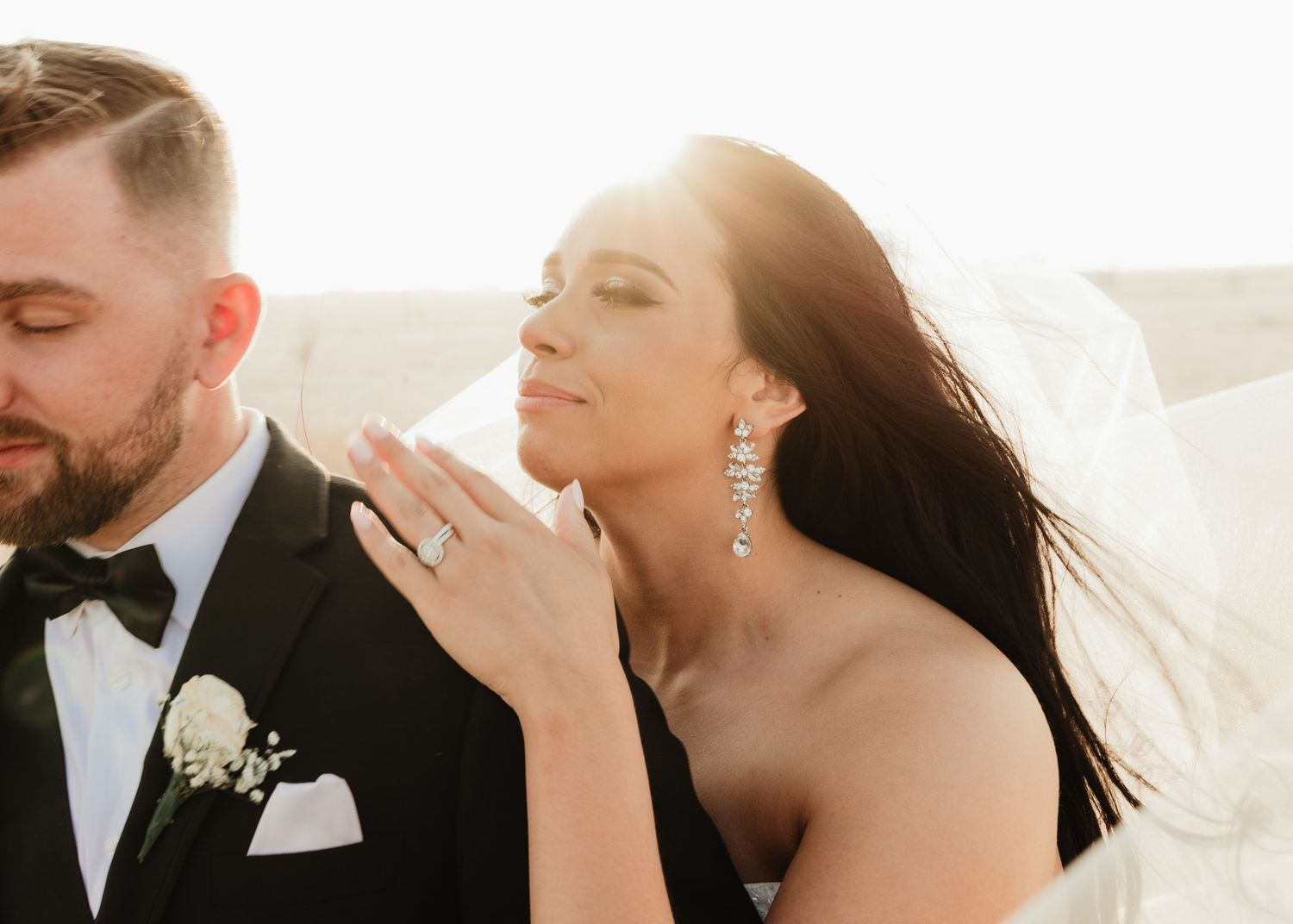 Bride and groom share romantic moment in golden sunlight, bride wearing diamond earrings and engagement ring, groom in black tuxedo.
