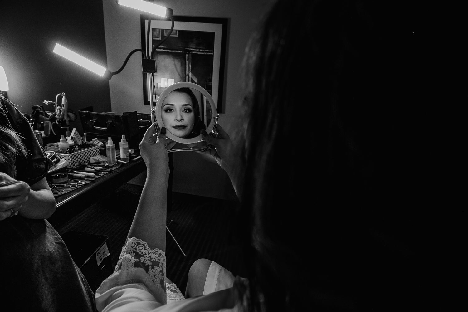 Bride getting makeup applied at a vanity mirror with bright lights in black and white.
