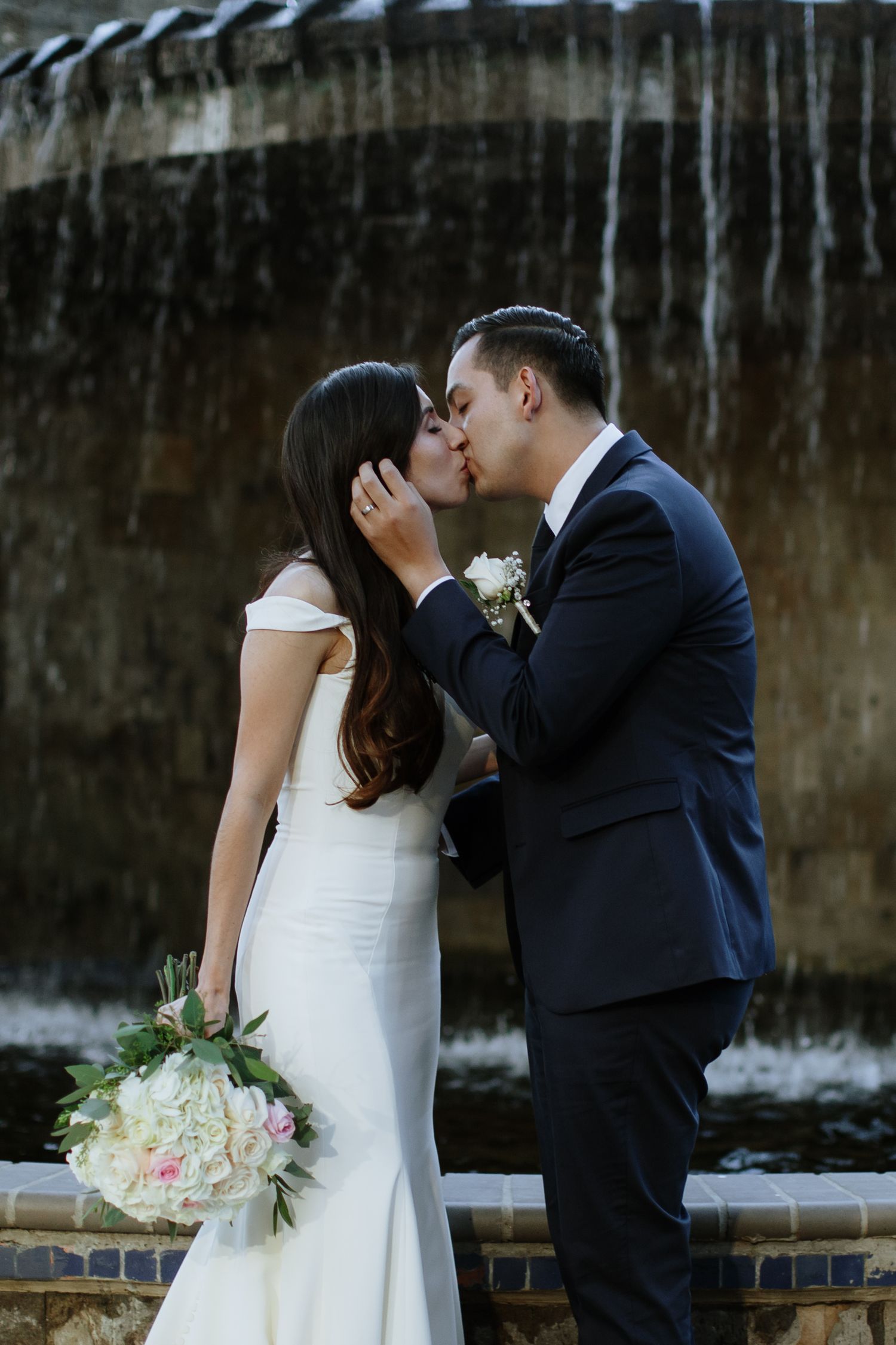 Bride and groom share romantic kiss in front of stone fountain, bride holding white and pink rose bouquet.