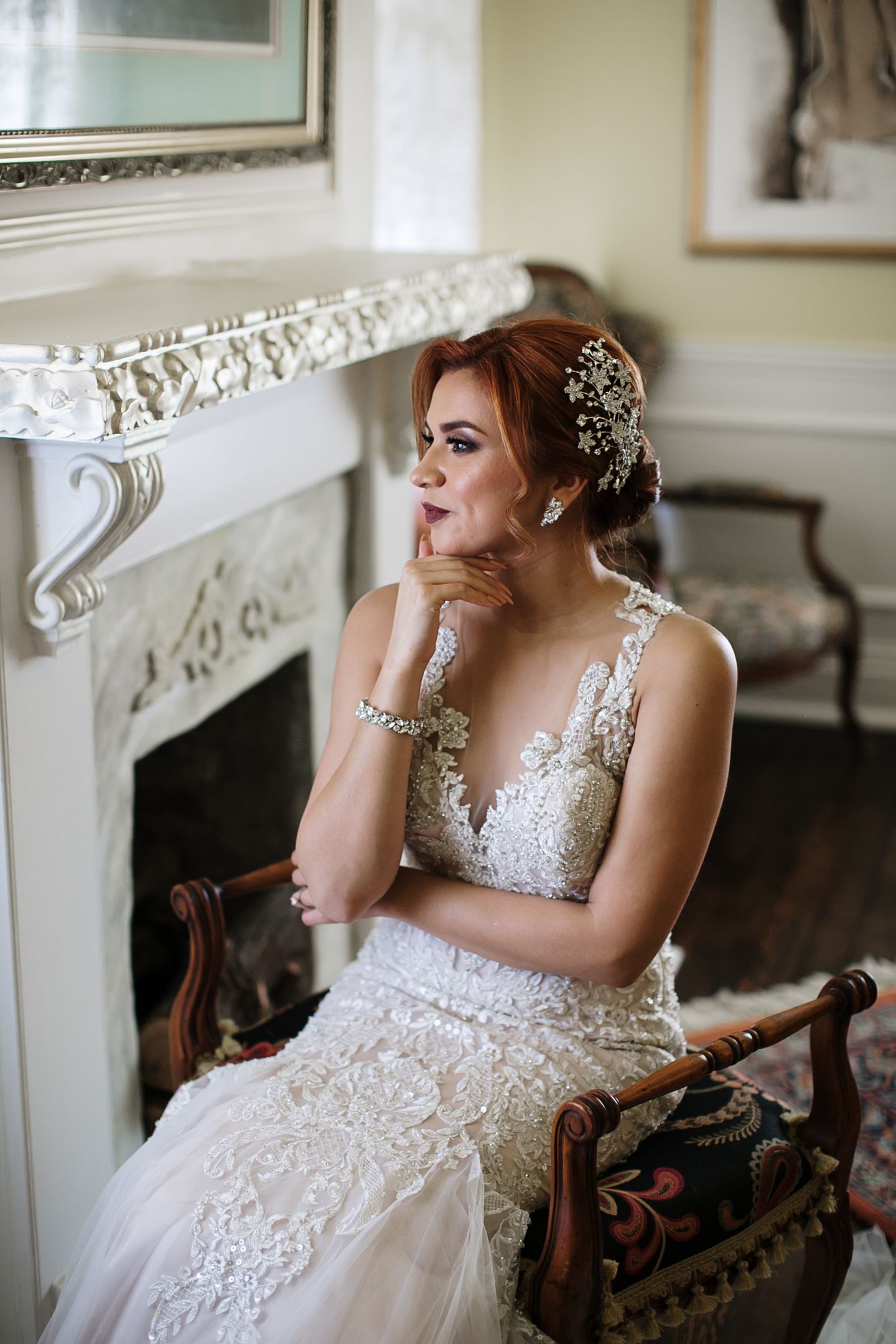 A bride in a lace wedding gown sits pensively by a white fireplace, wearing a floral hair piece and bracelet.