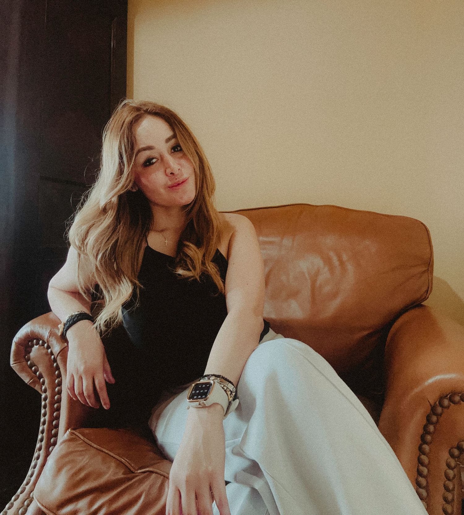 A person lounges casually on a brown leather armchair wearing a black top and white pants in warm lighting.