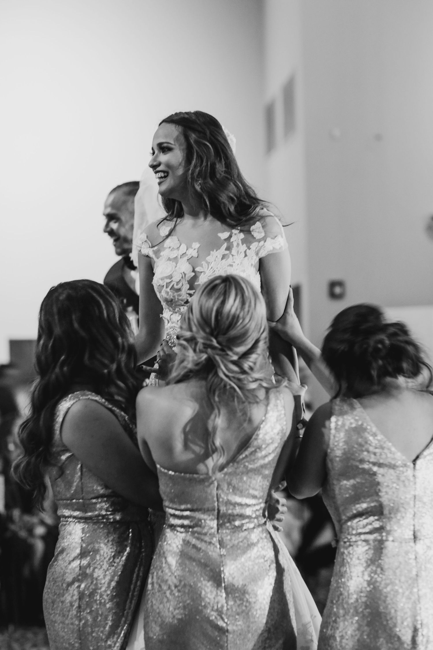 Bride in floral lace gown laughs joyfully while being lifted by bridesmaids in sequin dresses at wedding reception.
