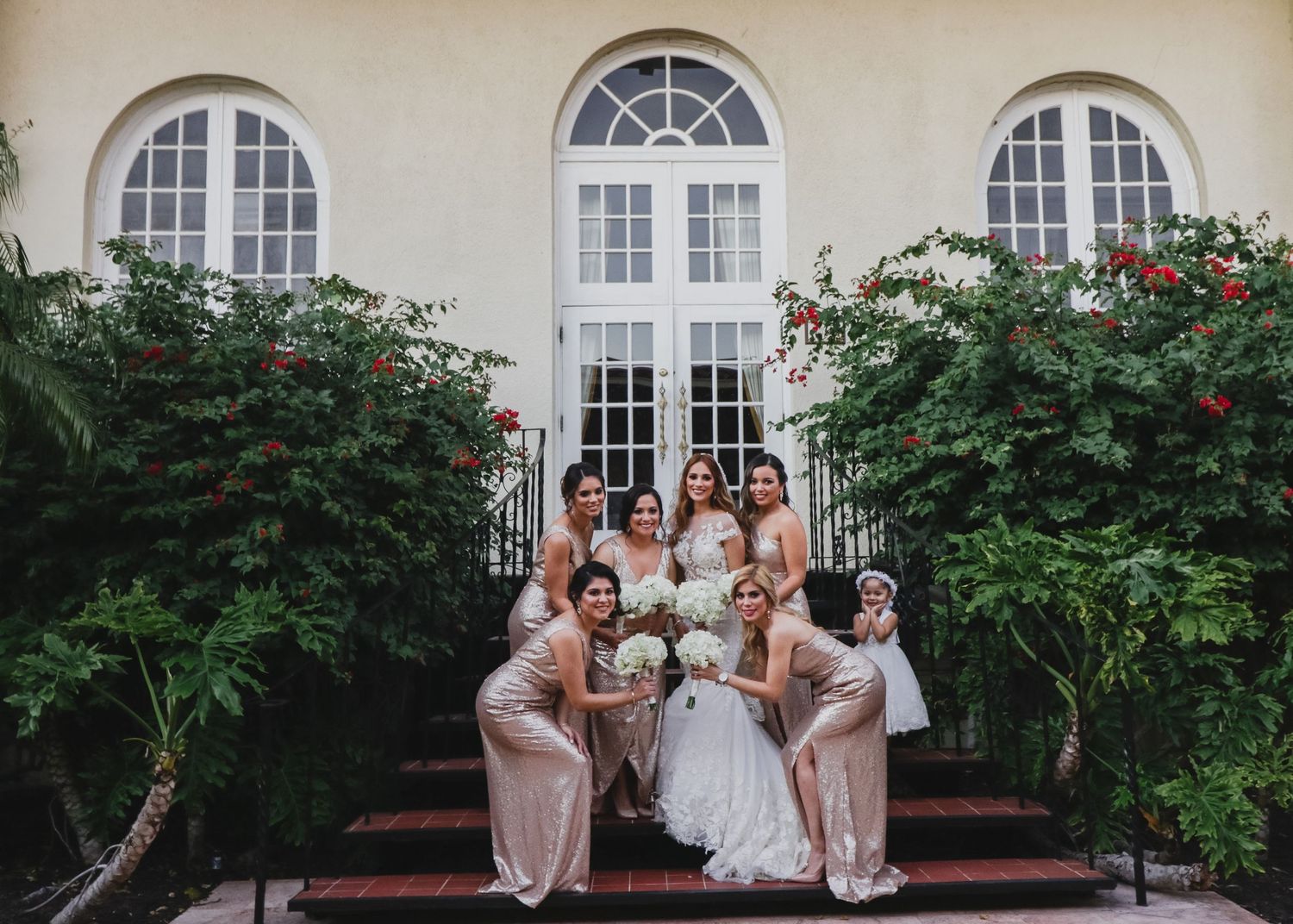 Bride in strapless white gown poses with seven bridesmaids in blush dresses on steps surrounded by roses.