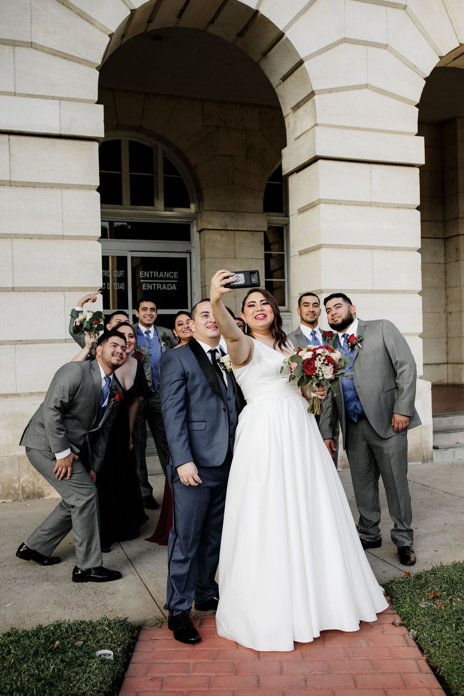 Bride taking a selfie with groom and groomsmen outside a building entrance on wedding day.