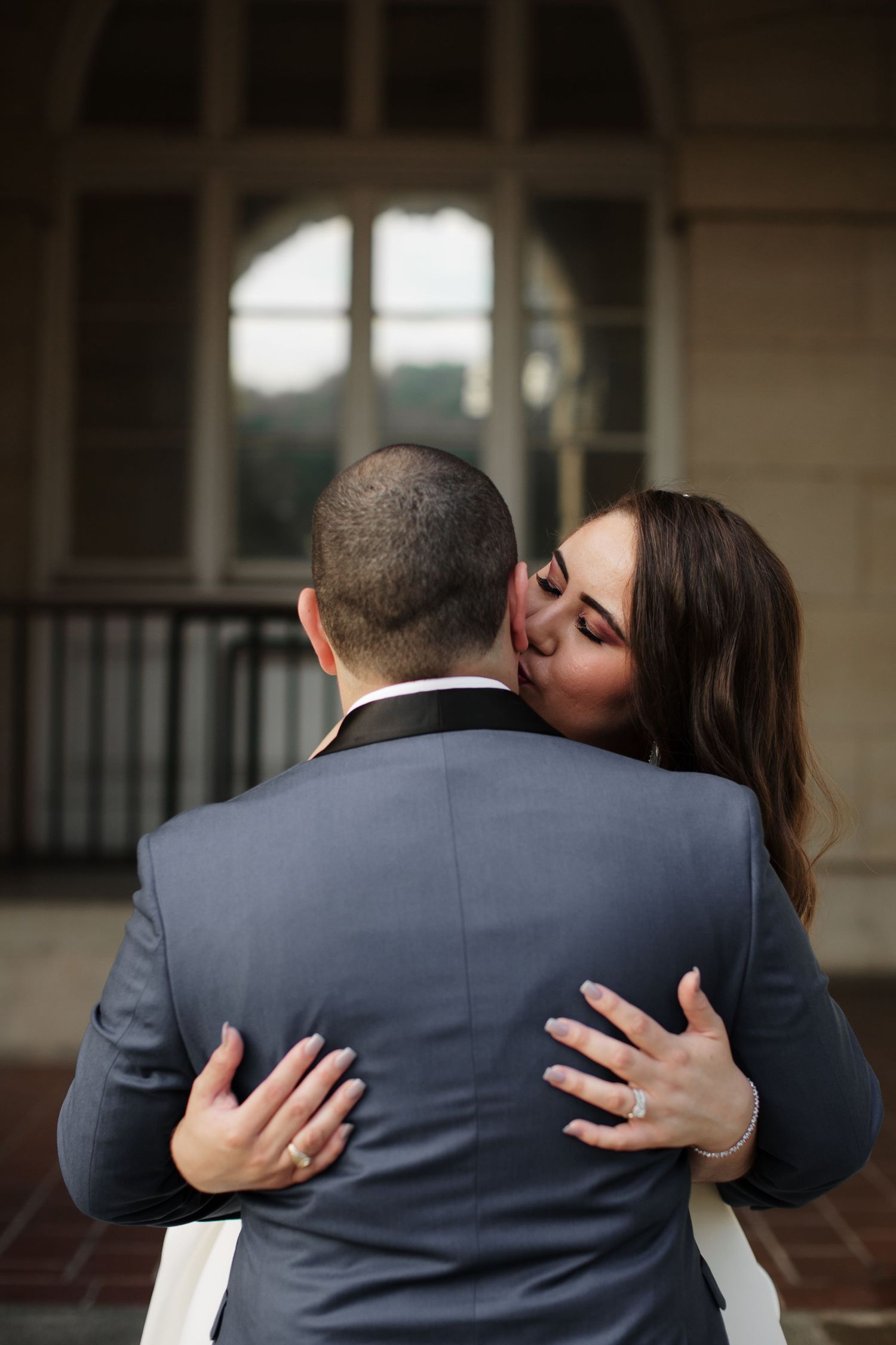 Bride in white dress embracing and kissing groom in gray suit from behind near arched window.