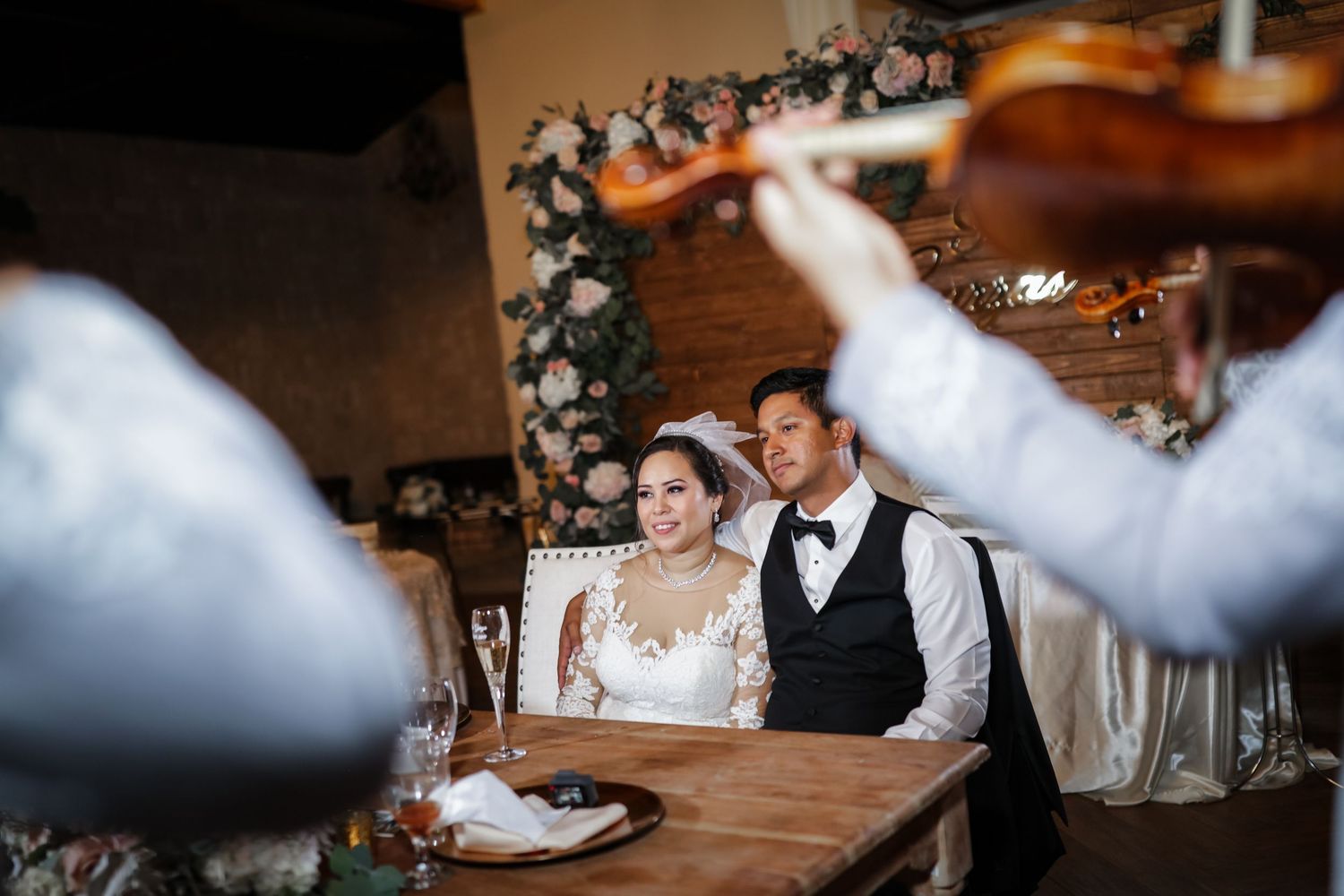 Bride and groom seated at reception table while violinists serenade them with live music.