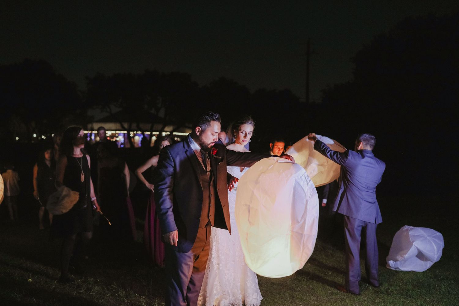 Wedding guests release glowing sky lanterns into the night sky during outdoor evening celebration.