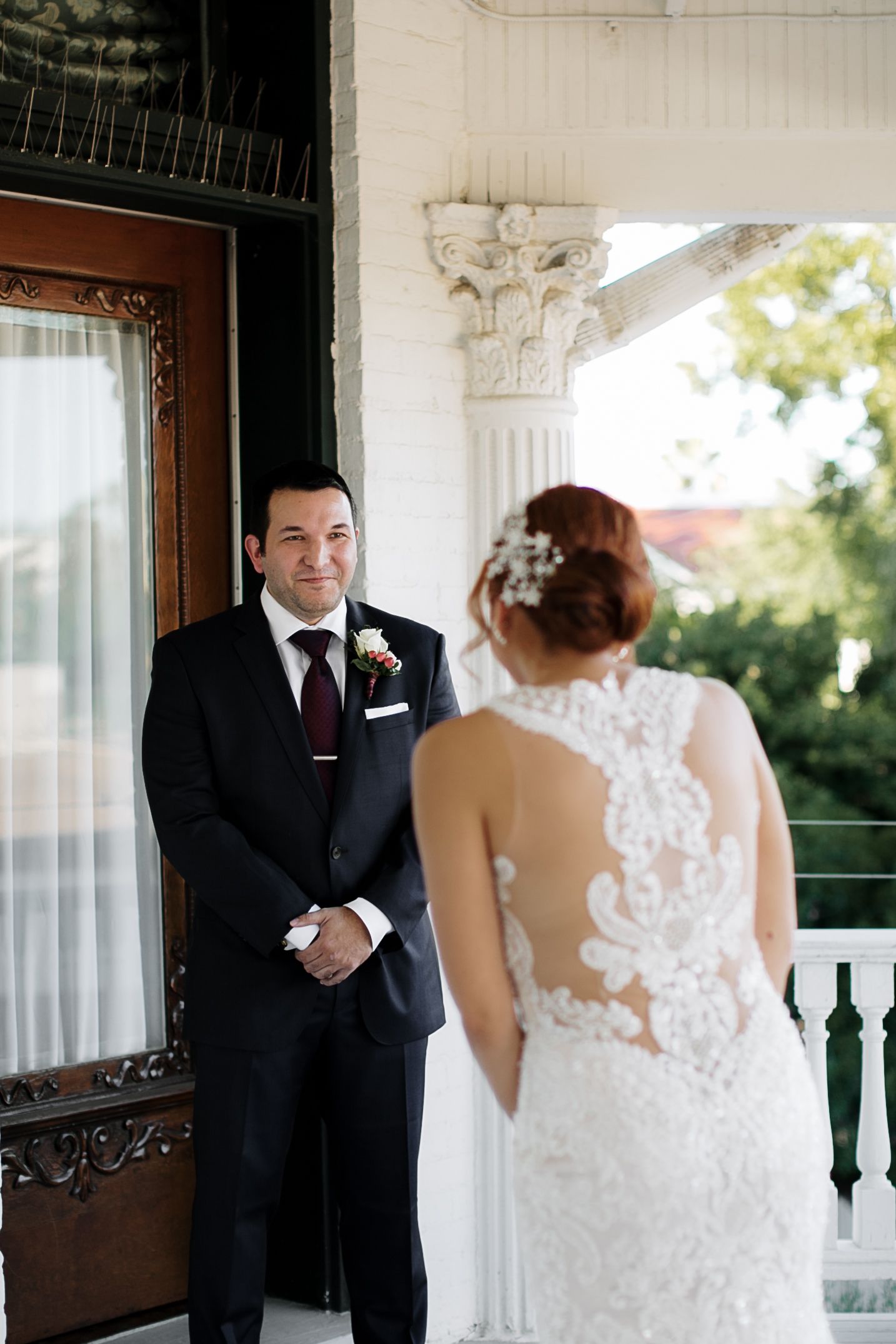 Groom in navy suit smiling at bride in lace backless wedding dress on porch.