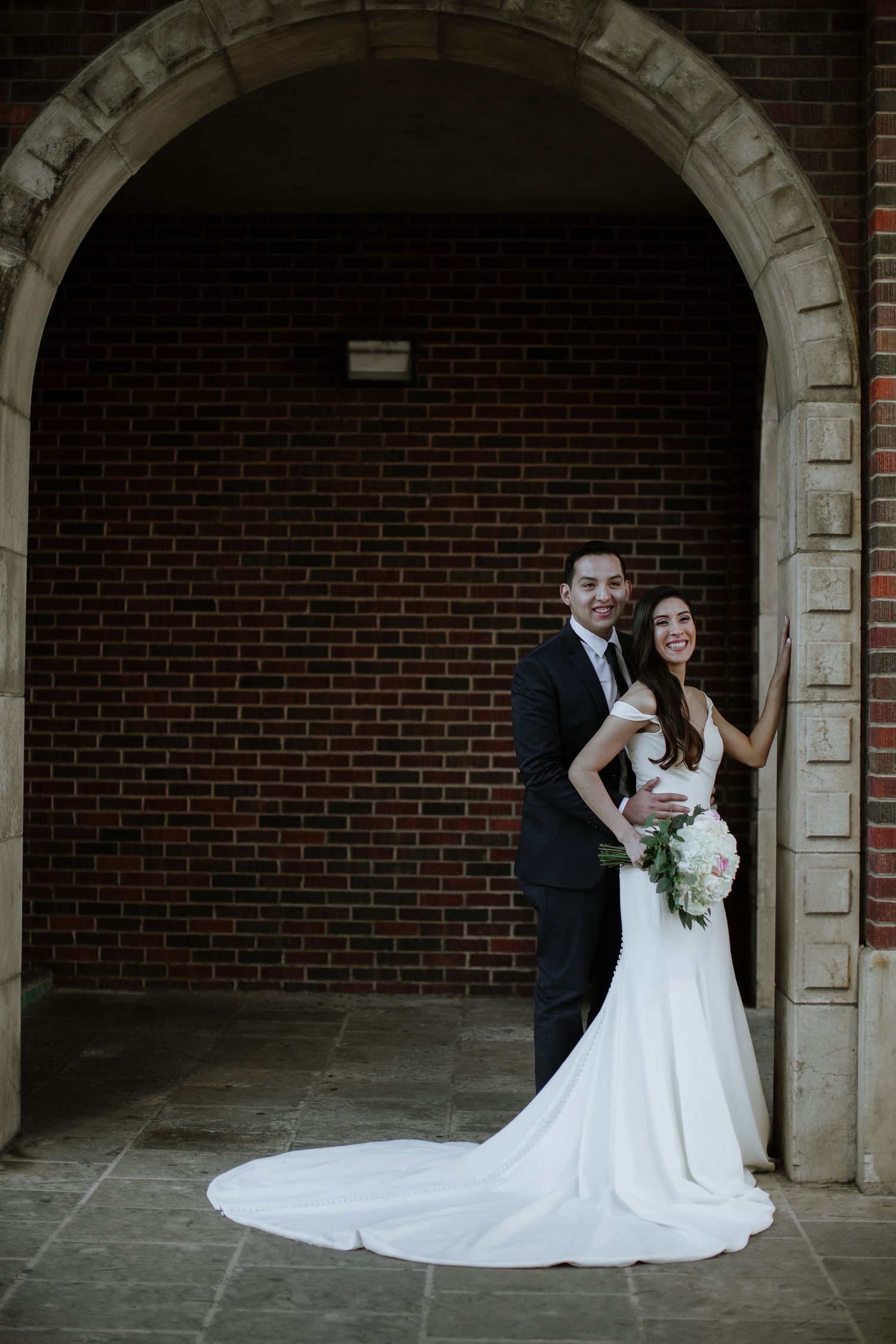 Bride and groom posing under a brick archway, bride holding a bouquet in a fitted white gown.