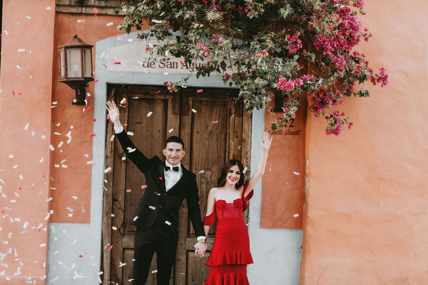 Happy couple celebrating with confetti outside colorful building, man in tuxedo and woman in red dress.