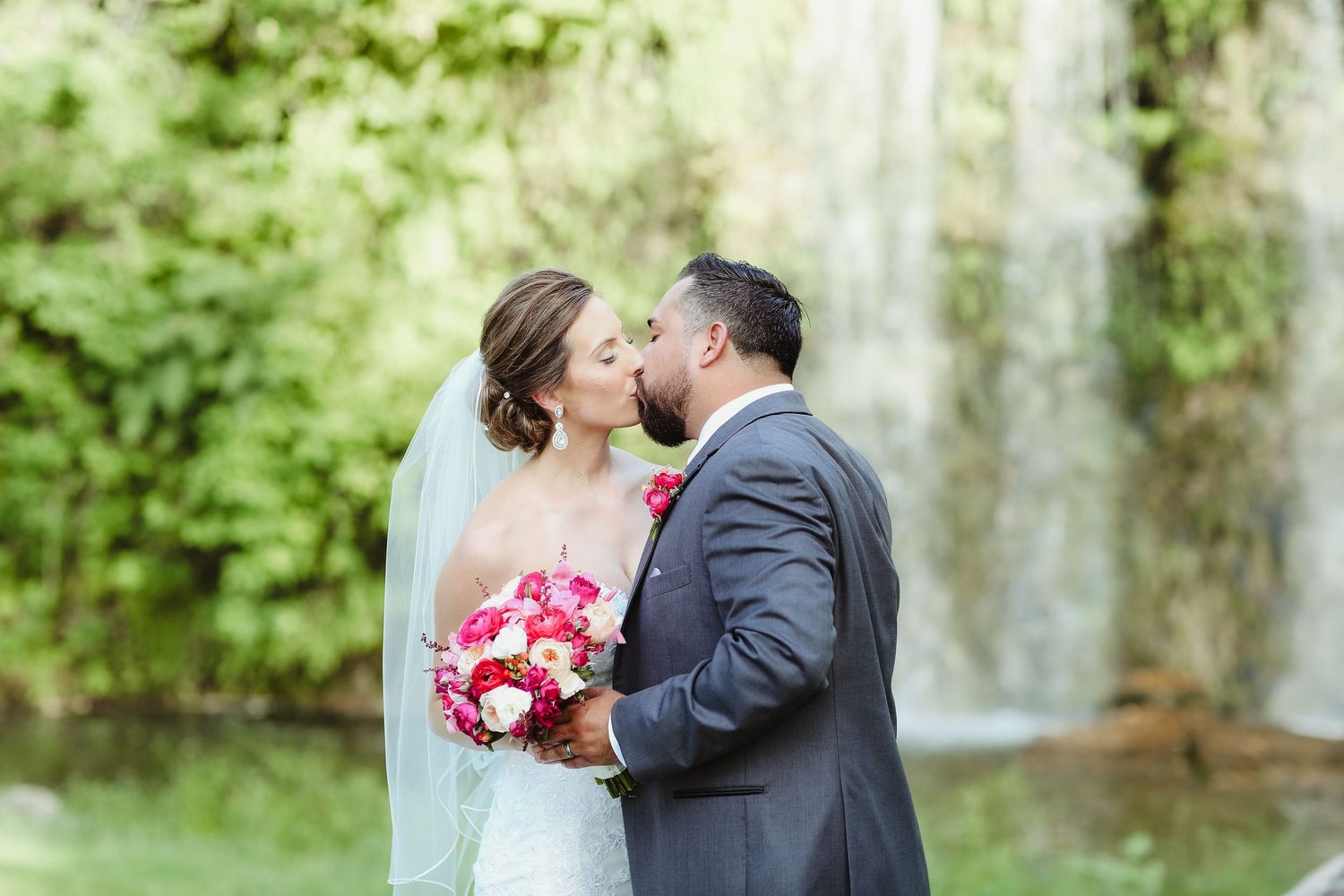 Bride and groom share a romantic kiss in front of a lush waterfall backdrop.
