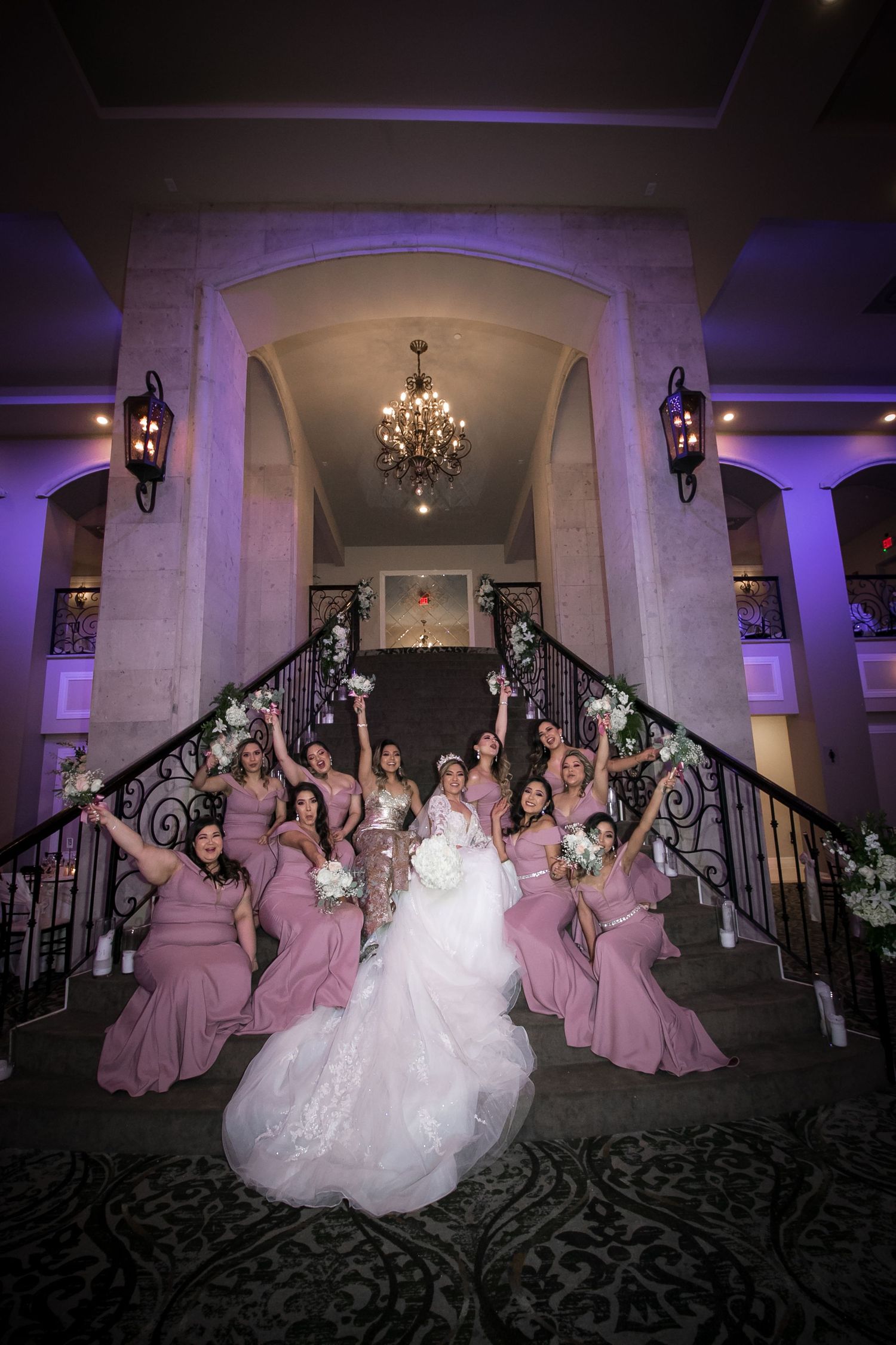 Bride and bridesmaids in mauve dresses celebrate with raised bouquets on elegant staircase.