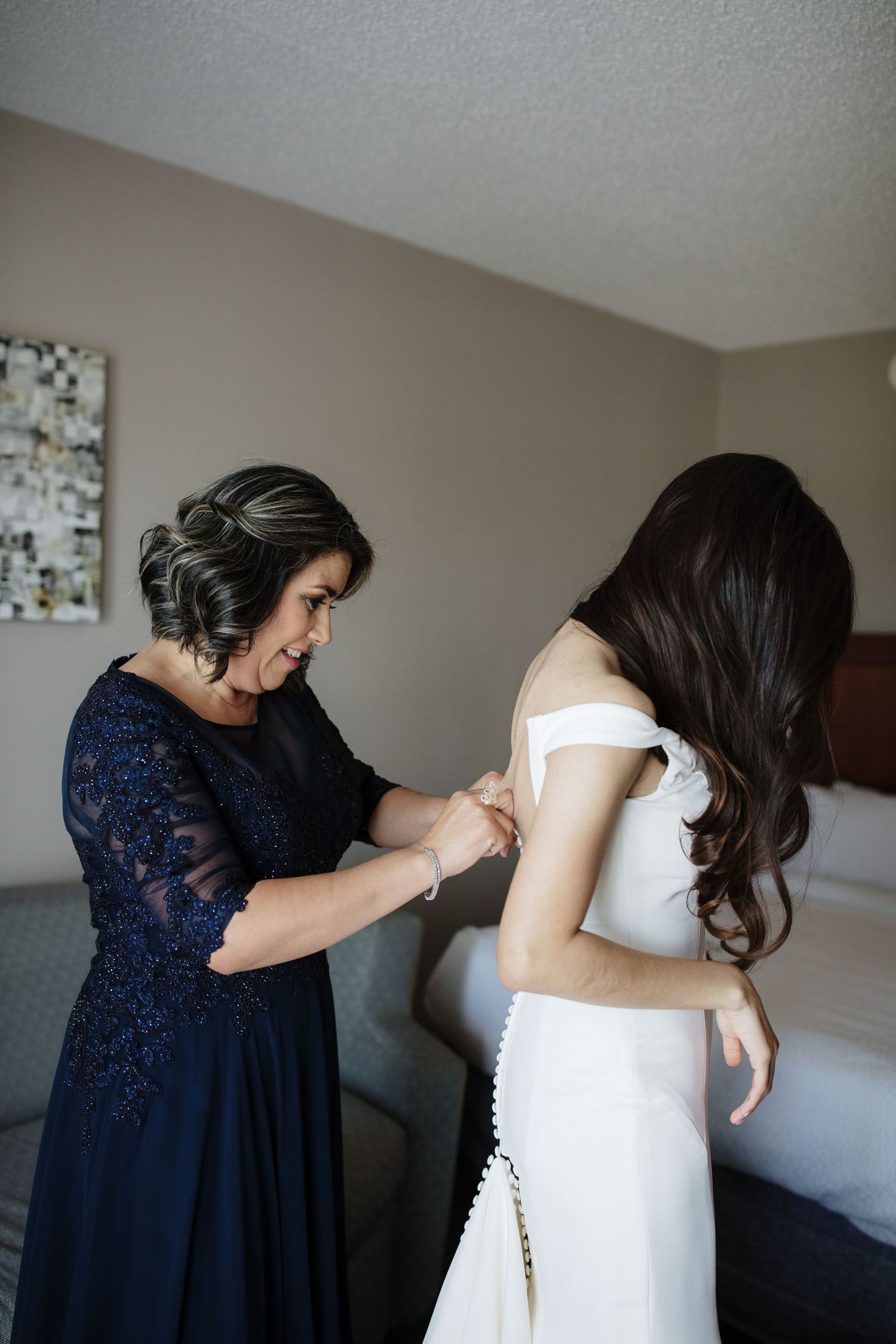 Mother helps bride fasten her off-shoulder white wedding gown in a warmly lit hotel room.
