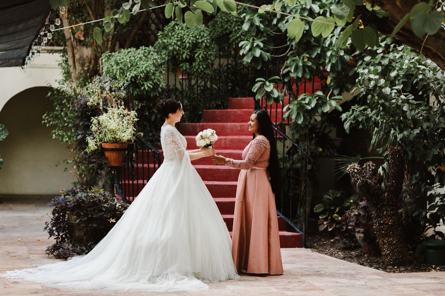 Bride in white gown receiving bouquet from bridesmaid on red staircase outdoors.