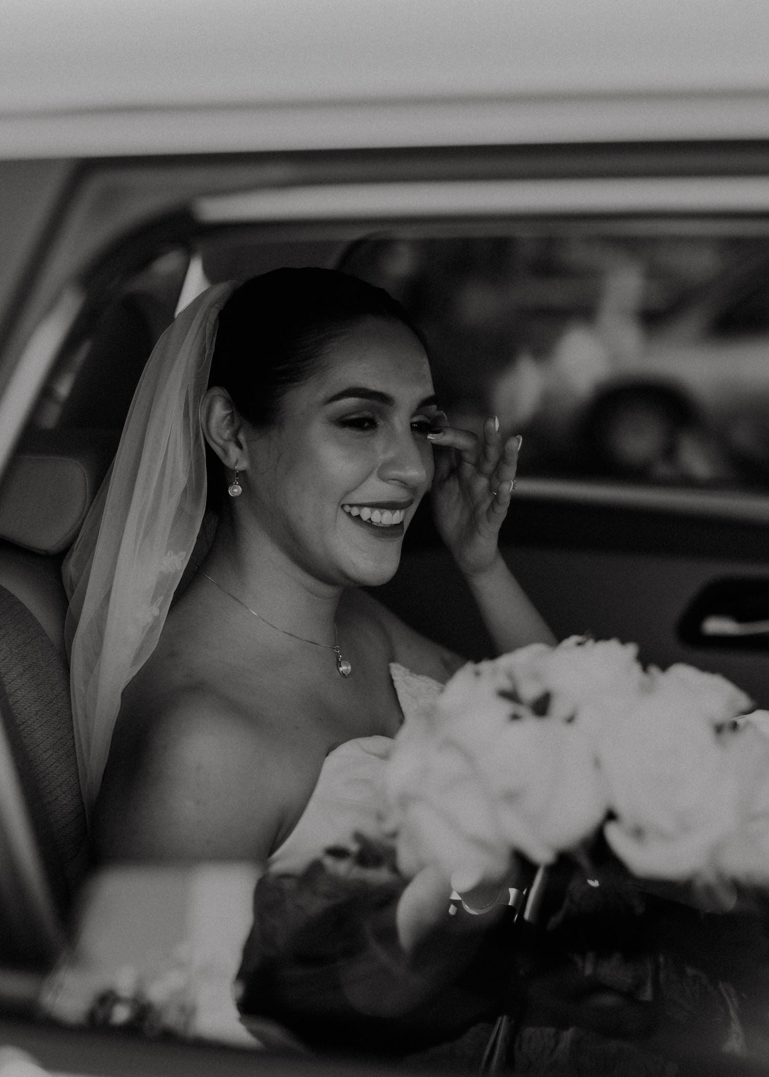 A joyful bride in a wedding veil holds a white bouquet while sitting in a car, wiping a happy tear.