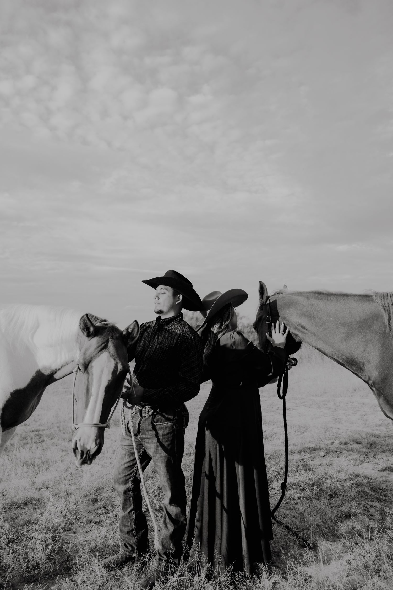 Black and white photo of a cowboy couple in hats standing with two horses in an open field.