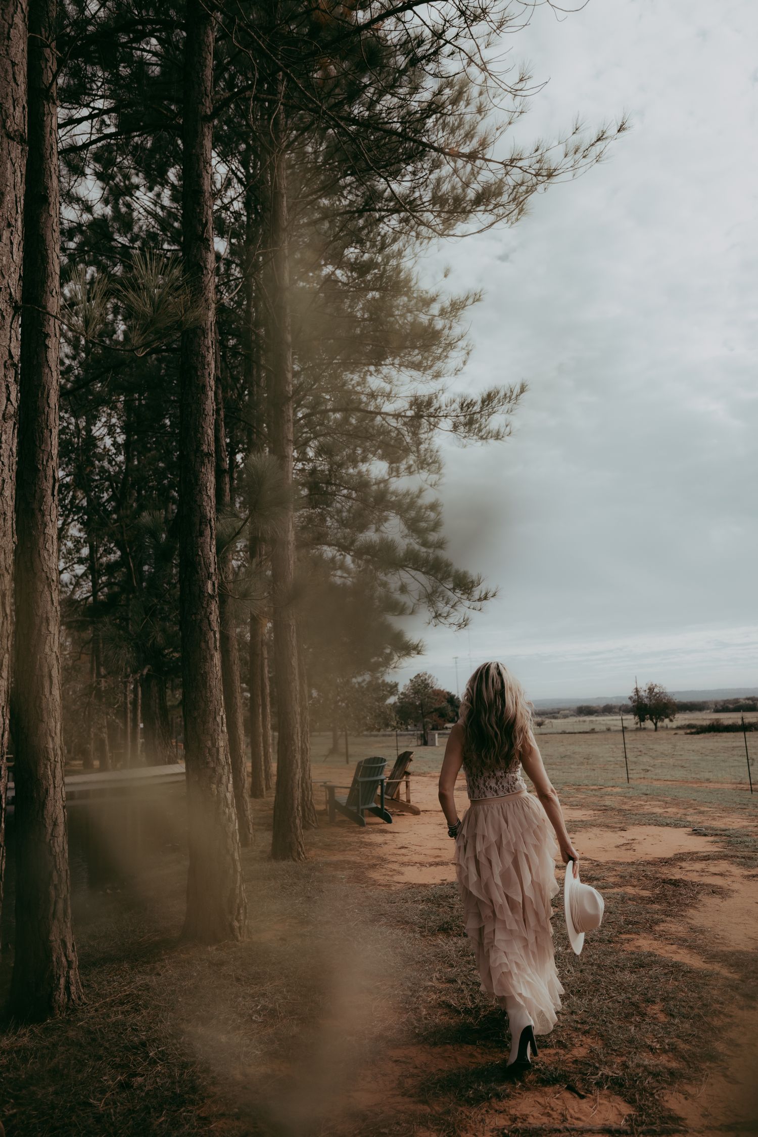 A dreamy photograph shows a feminine figure in a flowing dress walking along a pine-lined dirt path on a misty day.