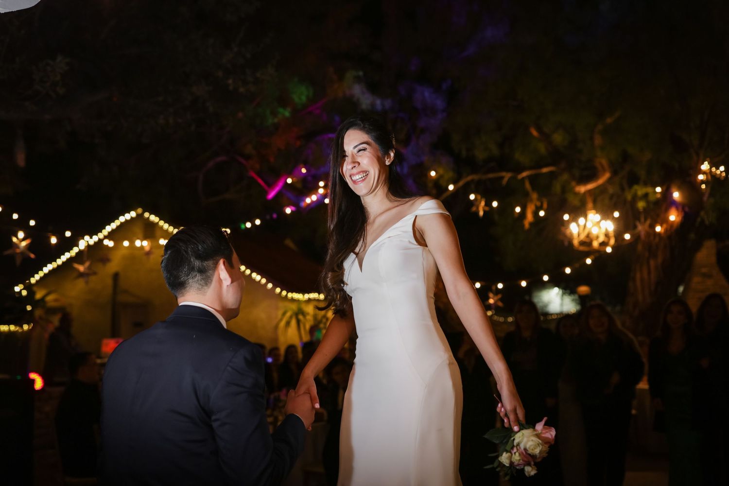 Smiling bride in white gown holding bouquet with groom under string lights at night wedding.